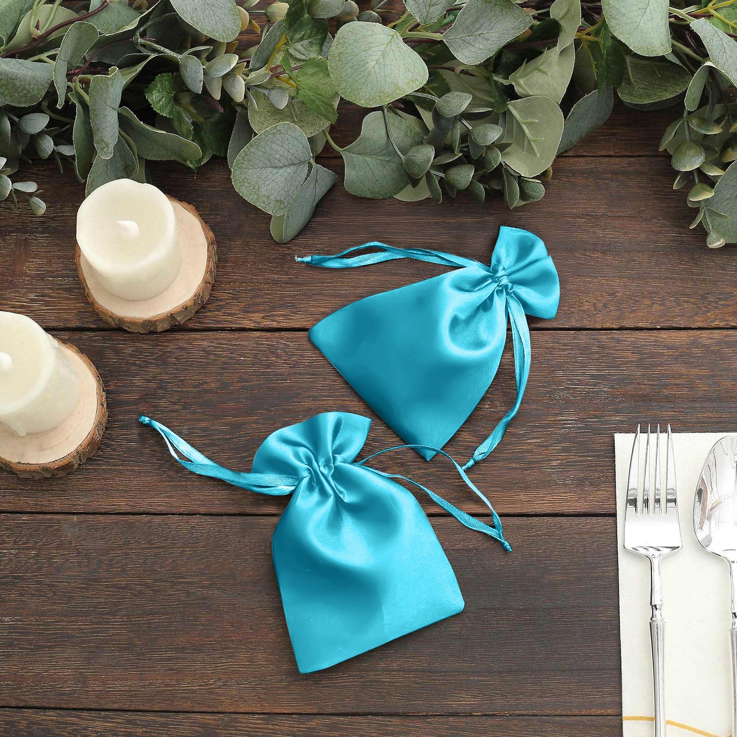 Turquoise satin drawstring favor bags on a wooden table with greenery.