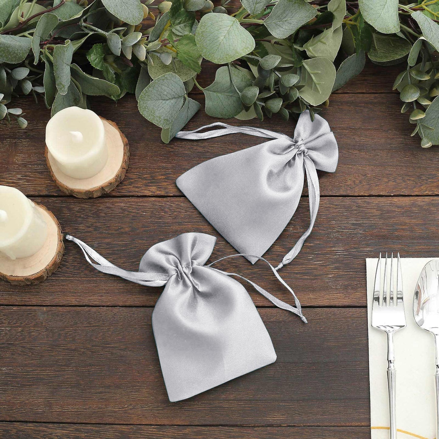 Silver satin drawstring pouch gift bags on a wooden table with greenery.
