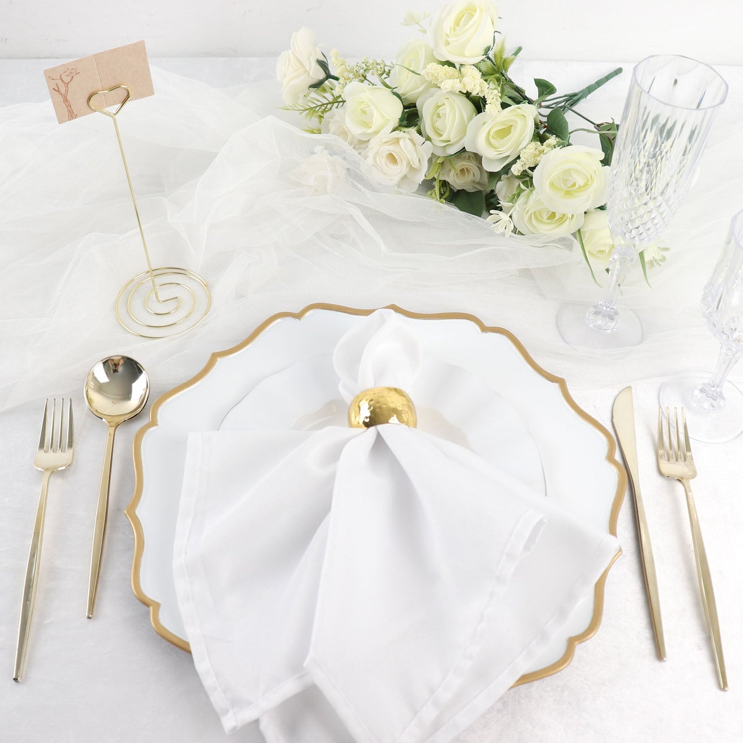 Gold metal heart card holder stand on an elegantly set table with white plates and flowers.