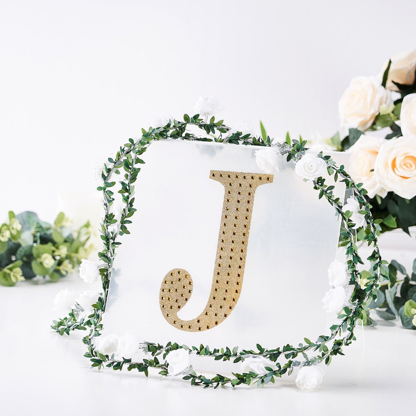 Gold decorative rhinestone letter "J" surrounded by greenery and flowers.