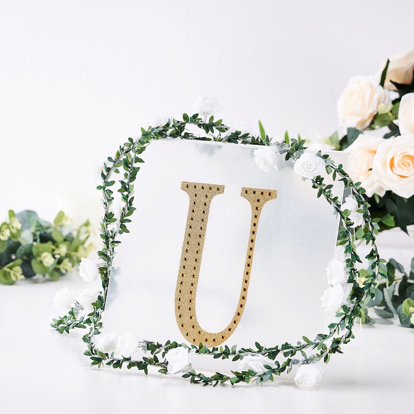 Gold decorative rhinestone letter "U" surrounded by greenery and flowers.