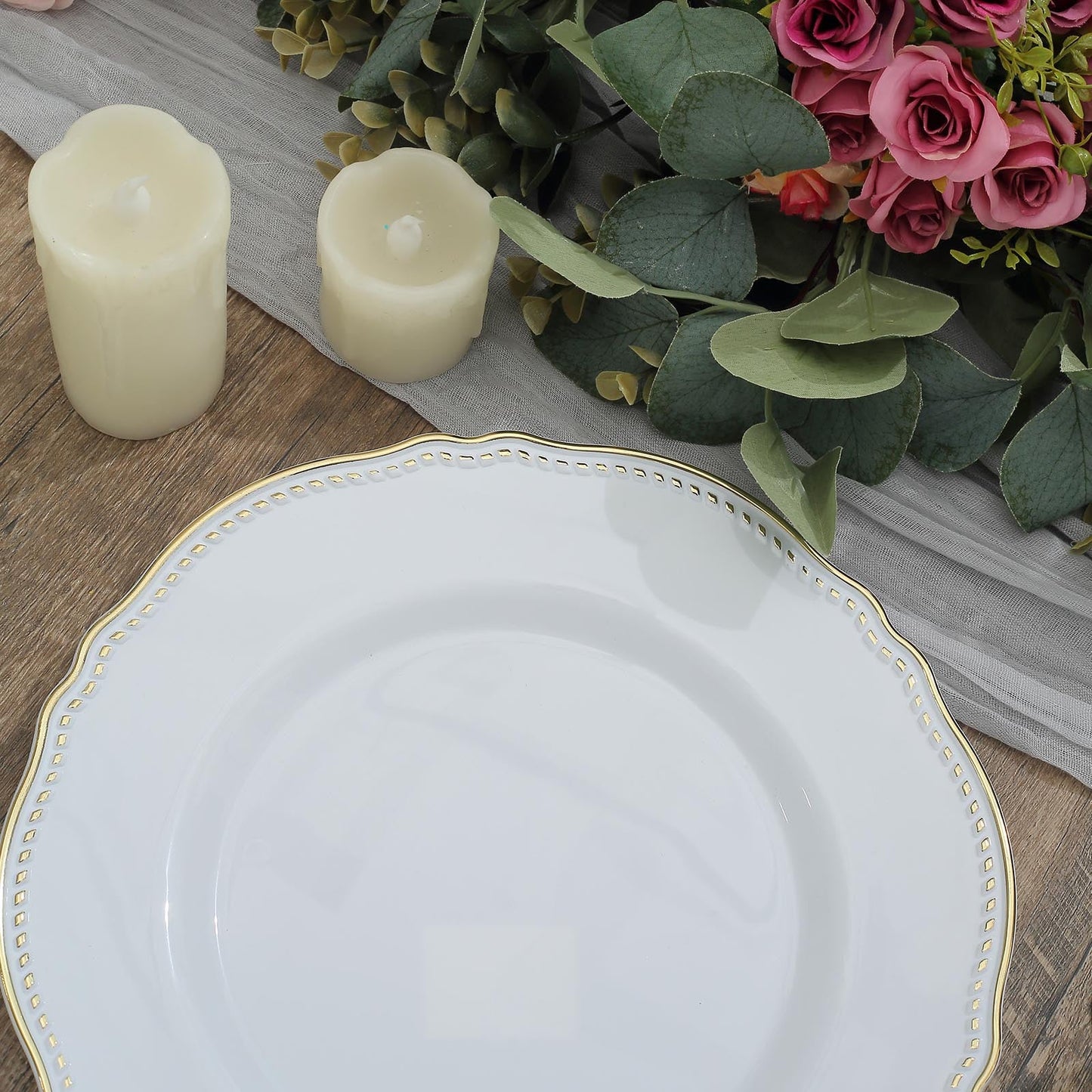 Elegant white plate with gold trim beside candles and floral arrangement on a wooden table.