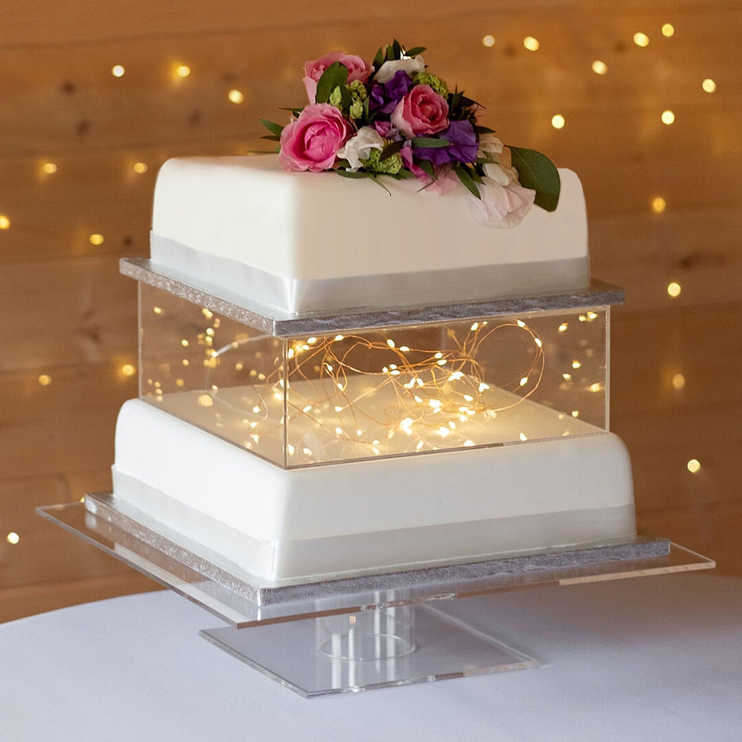 Clear acrylic cake box stand with illuminated display, featuring white cakes and flowers.