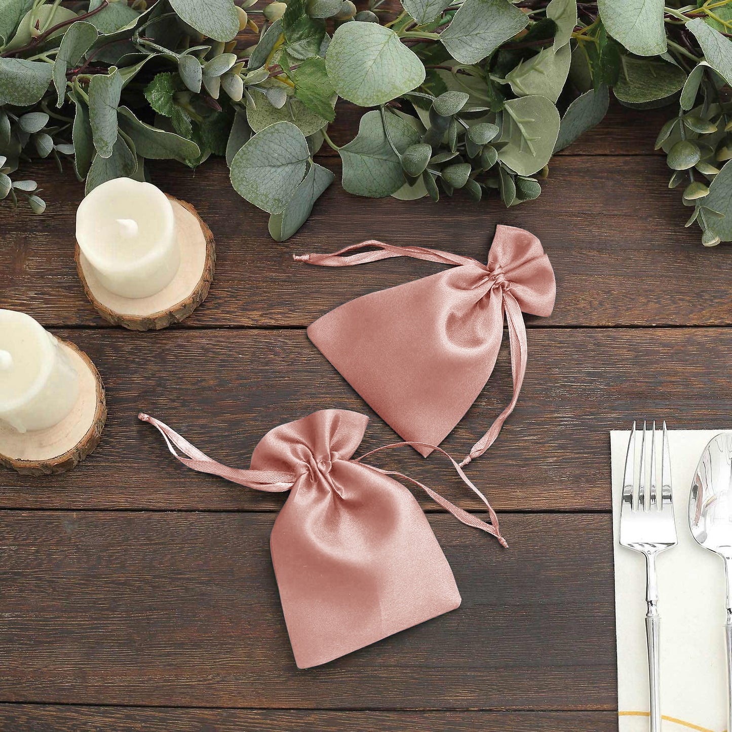 Dusty rose satin drawstring favor bags on a wooden table with greenery.