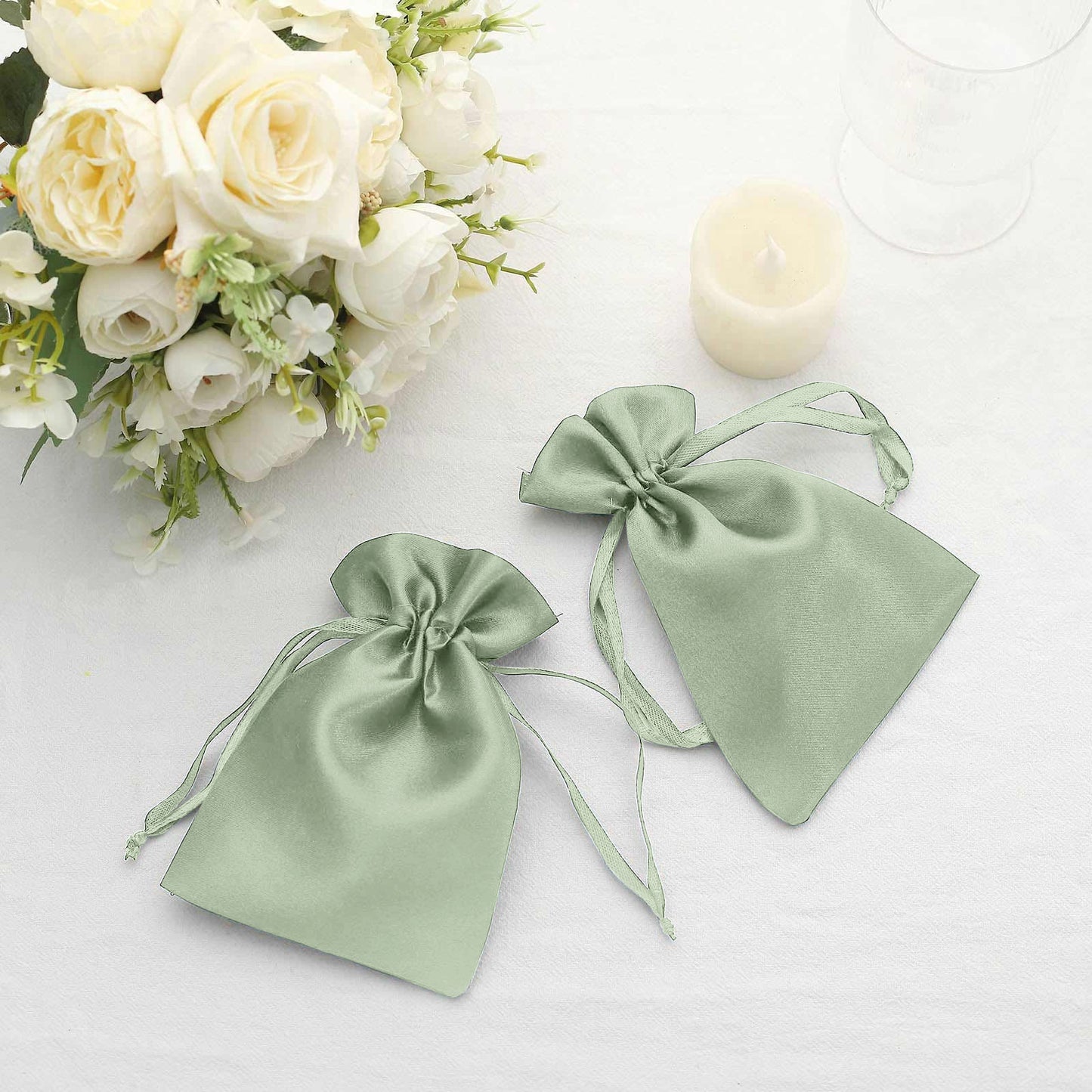 Sage green satin drawstring gift bags on a table with flowers.