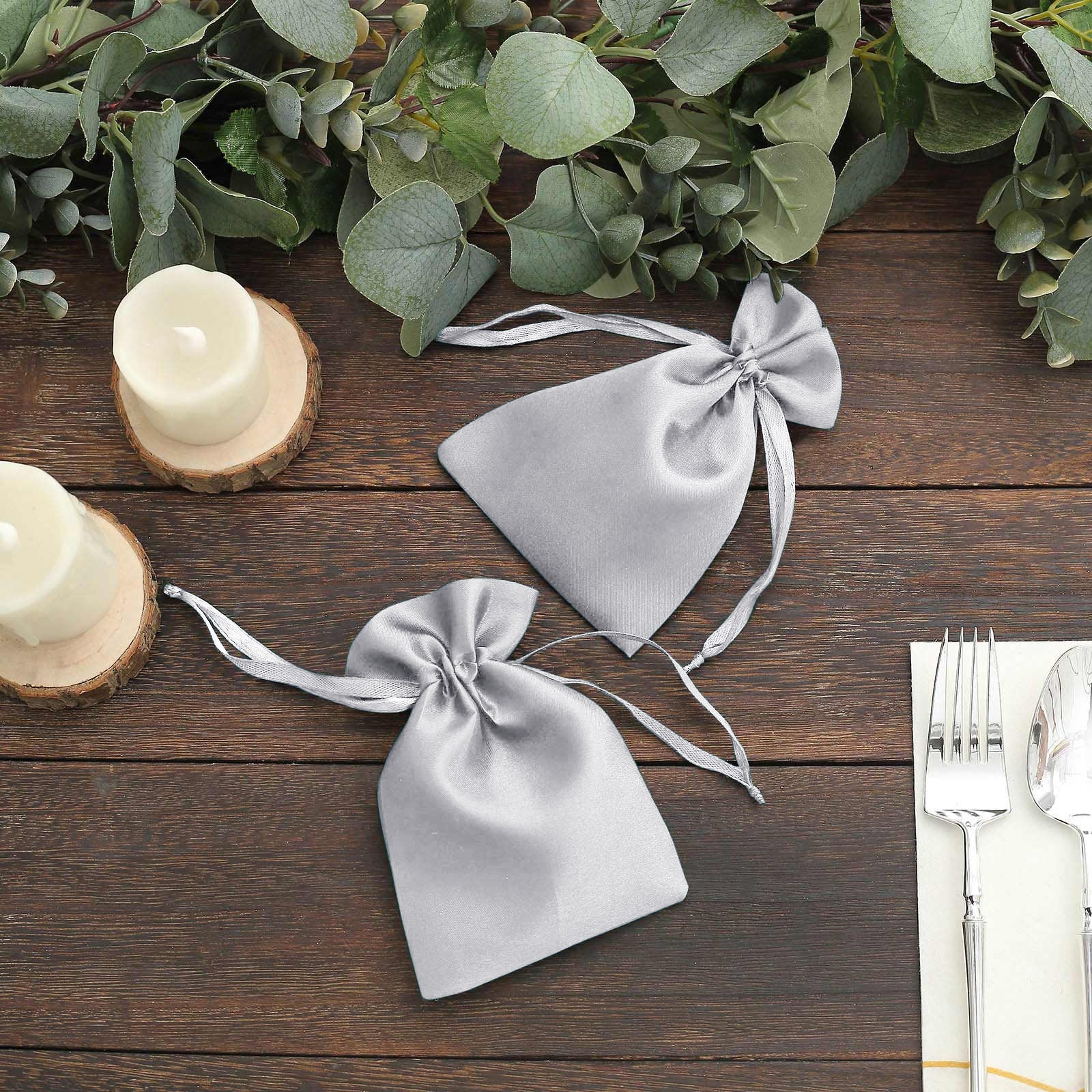 Silver satin drawstring pouch gift bags on a wooden table with greenery.