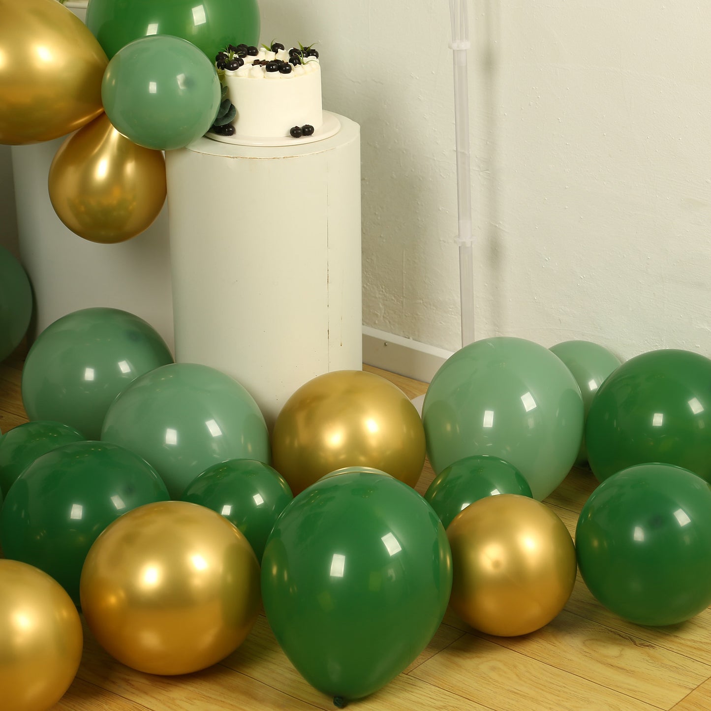 Gold and green biodegradable balloon arch kit on a floor with a cake.