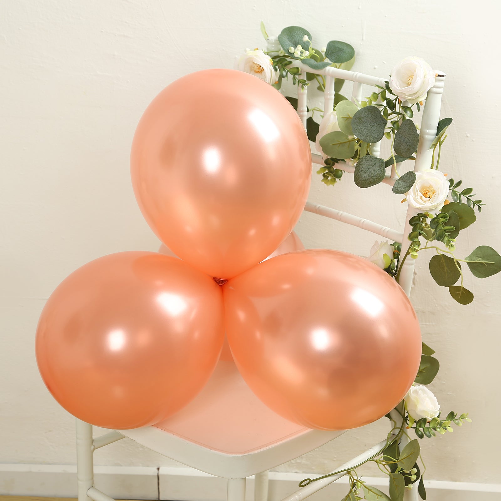 Rose gold biodegradable balloons arranged with greenery and white flowers.