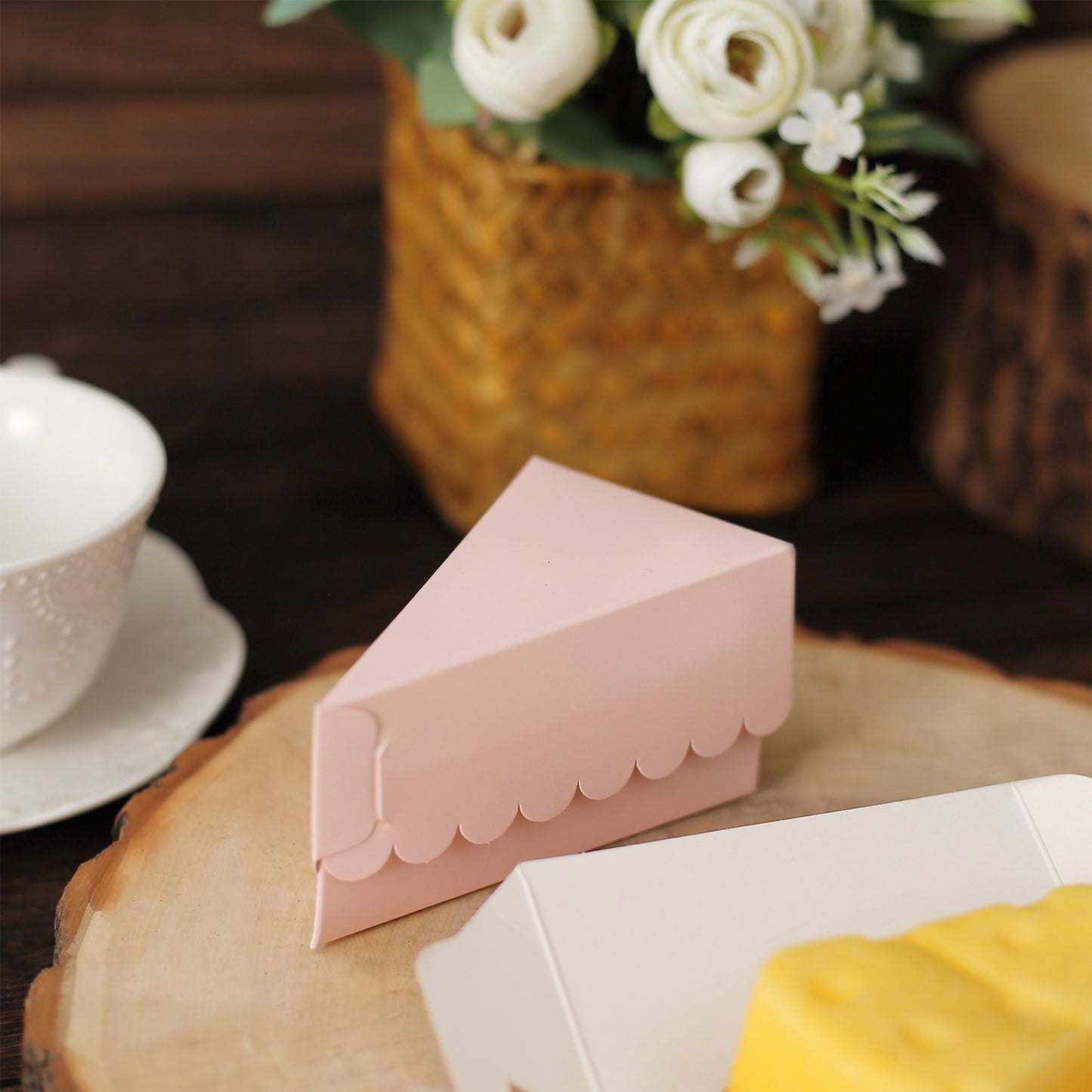 Blush triangular paper cake box on a wooden platter with flowers in the background.