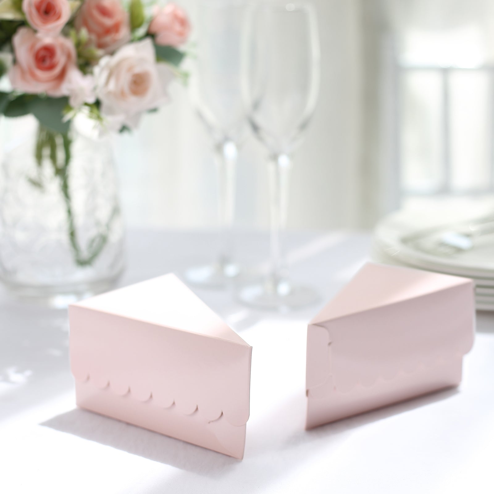 Blush triangular paper cake boxes on a table with flowers and glassware.