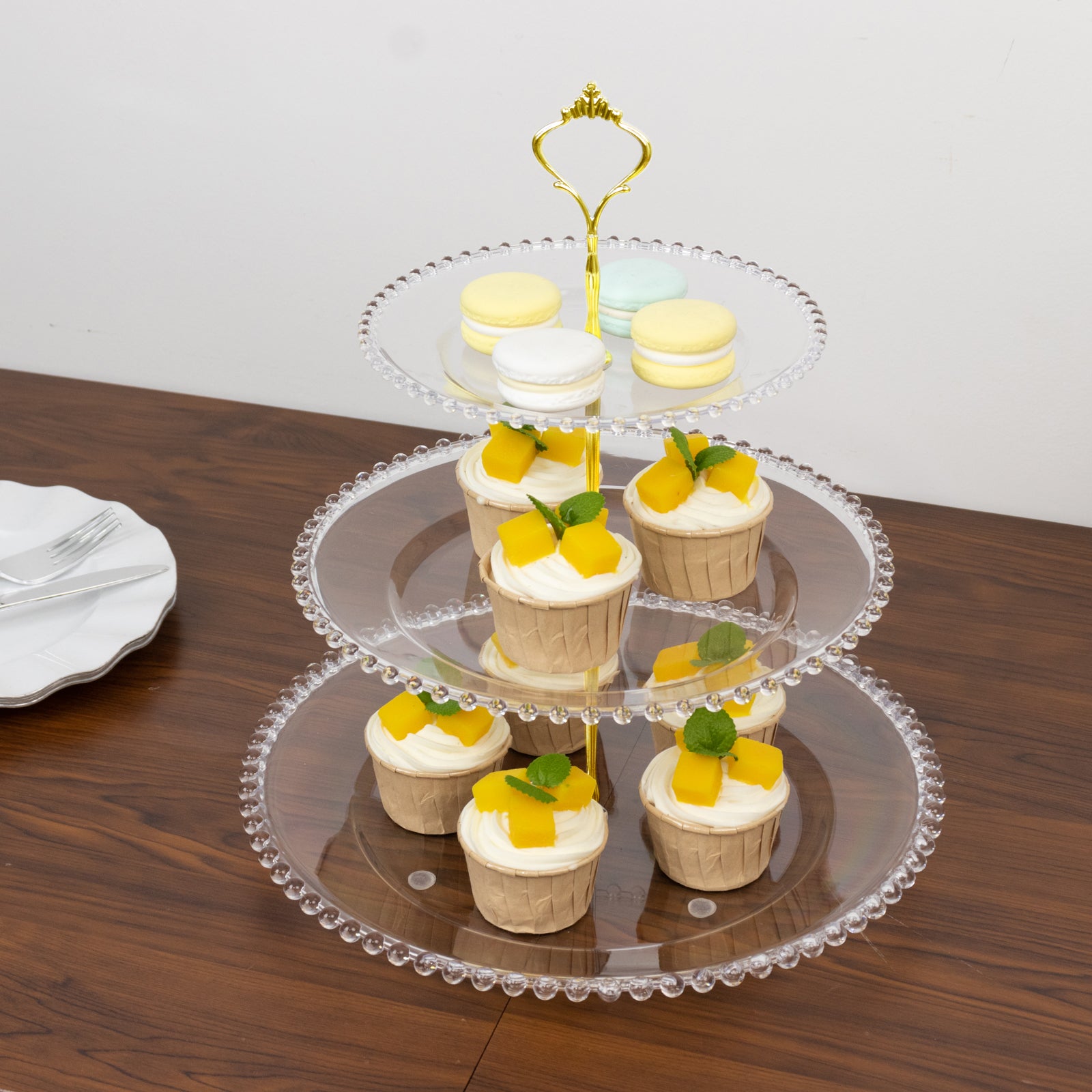 Clear 3-tier dessert display stand with cupcakes and macarons on a wooden table.