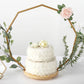 Gold floral hoop cake stand with wooden base and two-tiered white cake.