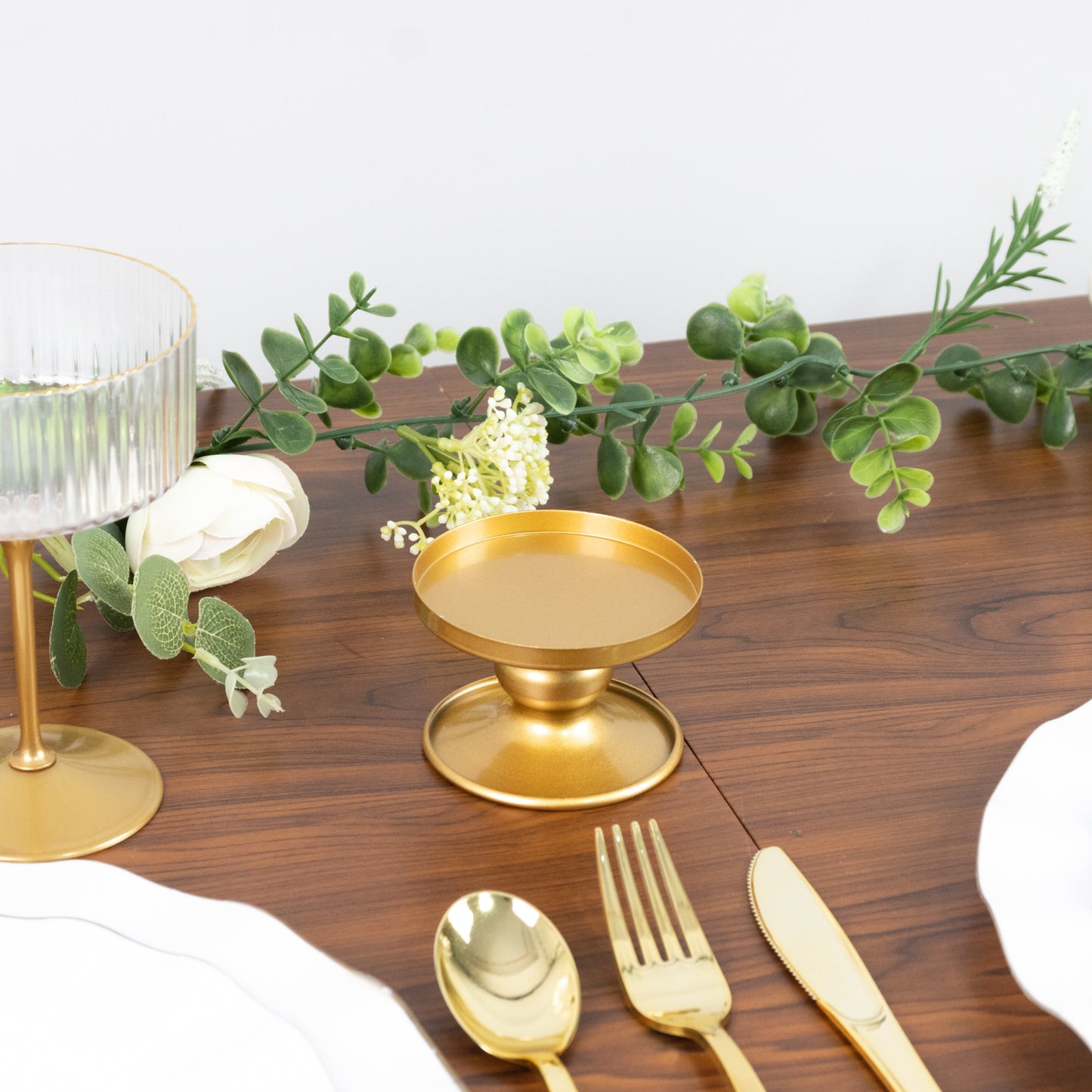 Gold metal candle holders set on a wooden table with greenery and tableware.