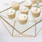 Gold geometric cake stand with square glass top displaying cupcakes.