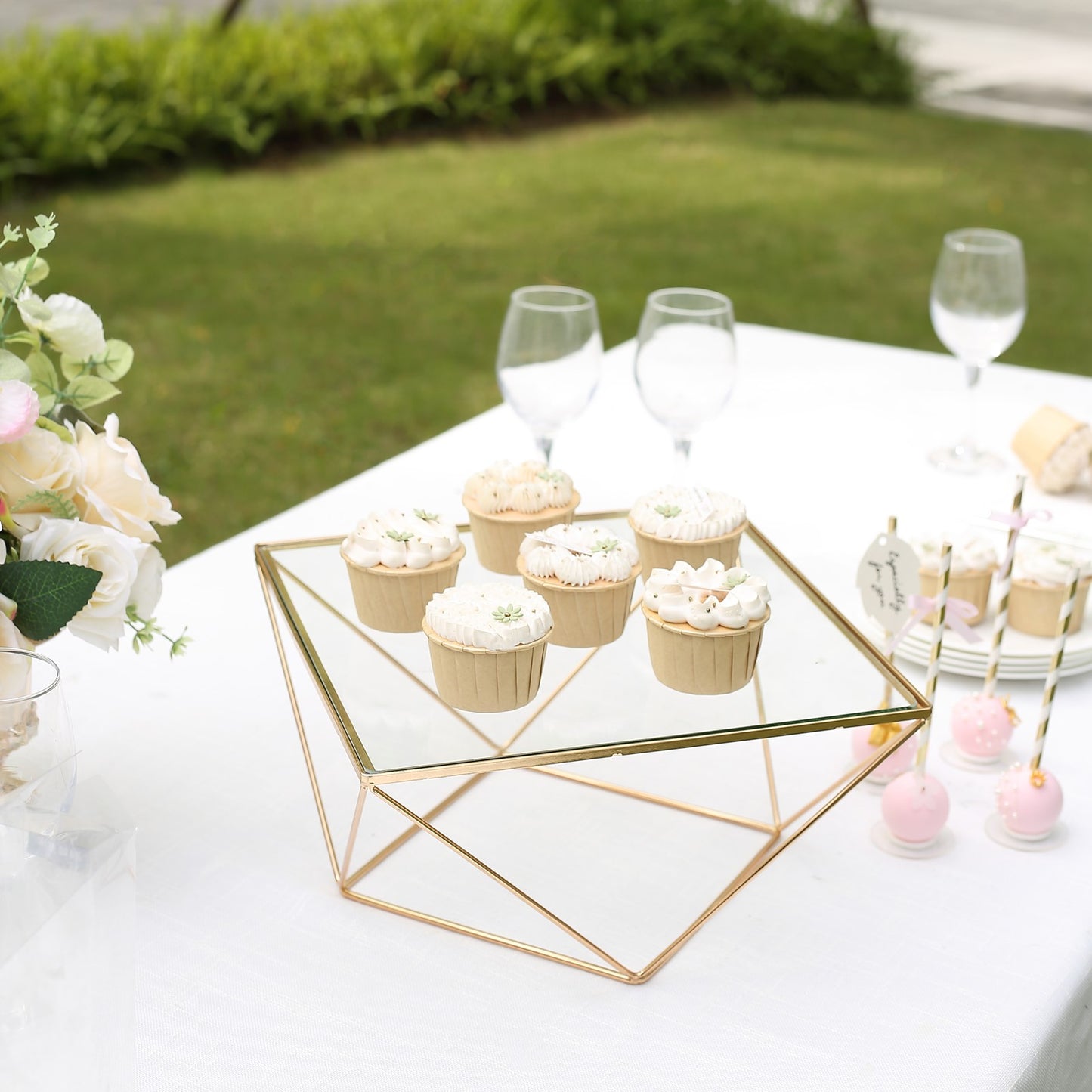 Gold geometric cake stand with square glass top displaying cupcakes.