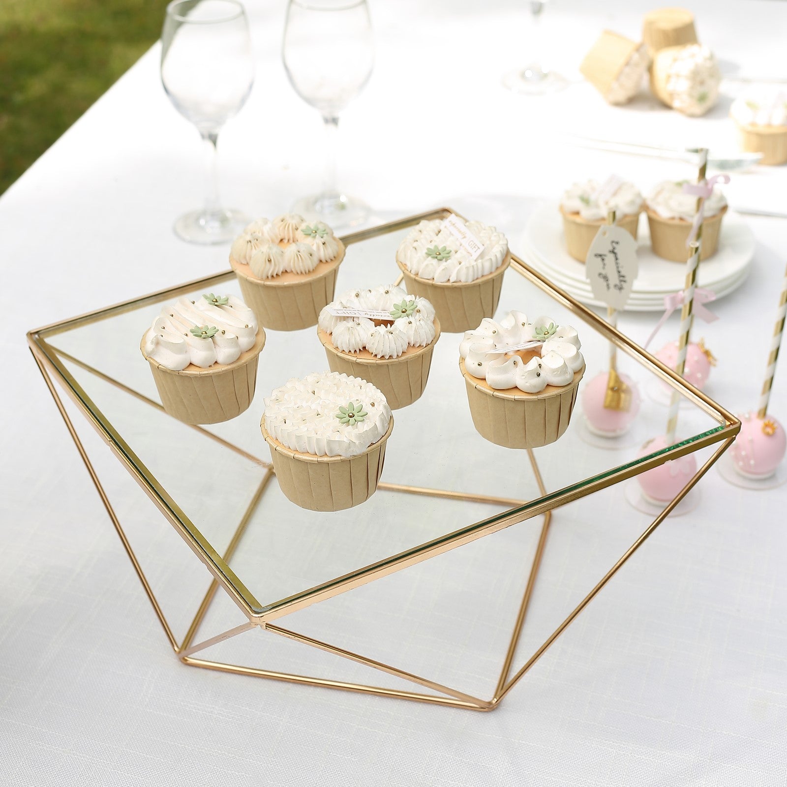 Gold geometric cake stand display with square glass top and cupcakes.