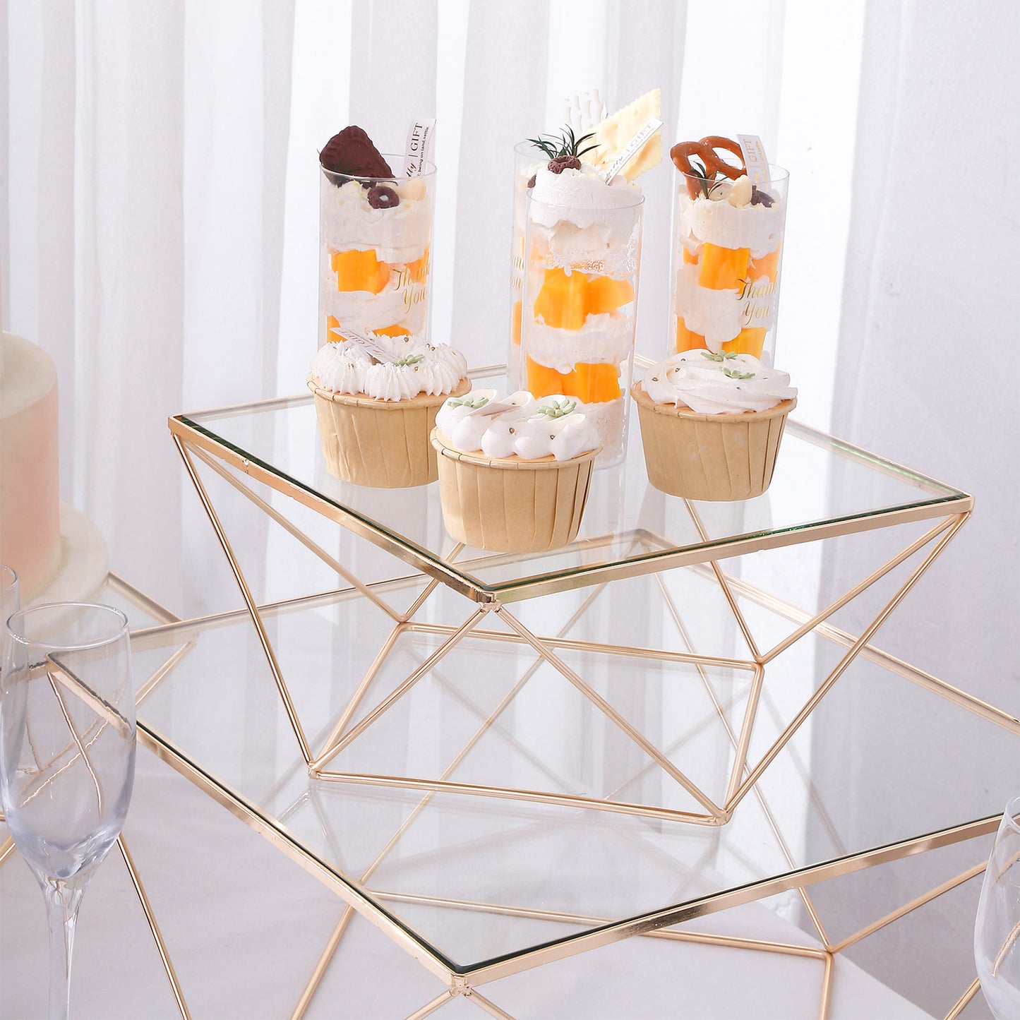 Gold metal geometric cake stand with square glass top displaying desserts.