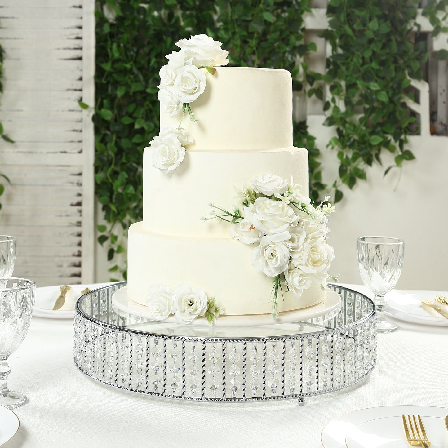 Silver round cake stand with crystal beads and glass top, featuring a white floral cake.