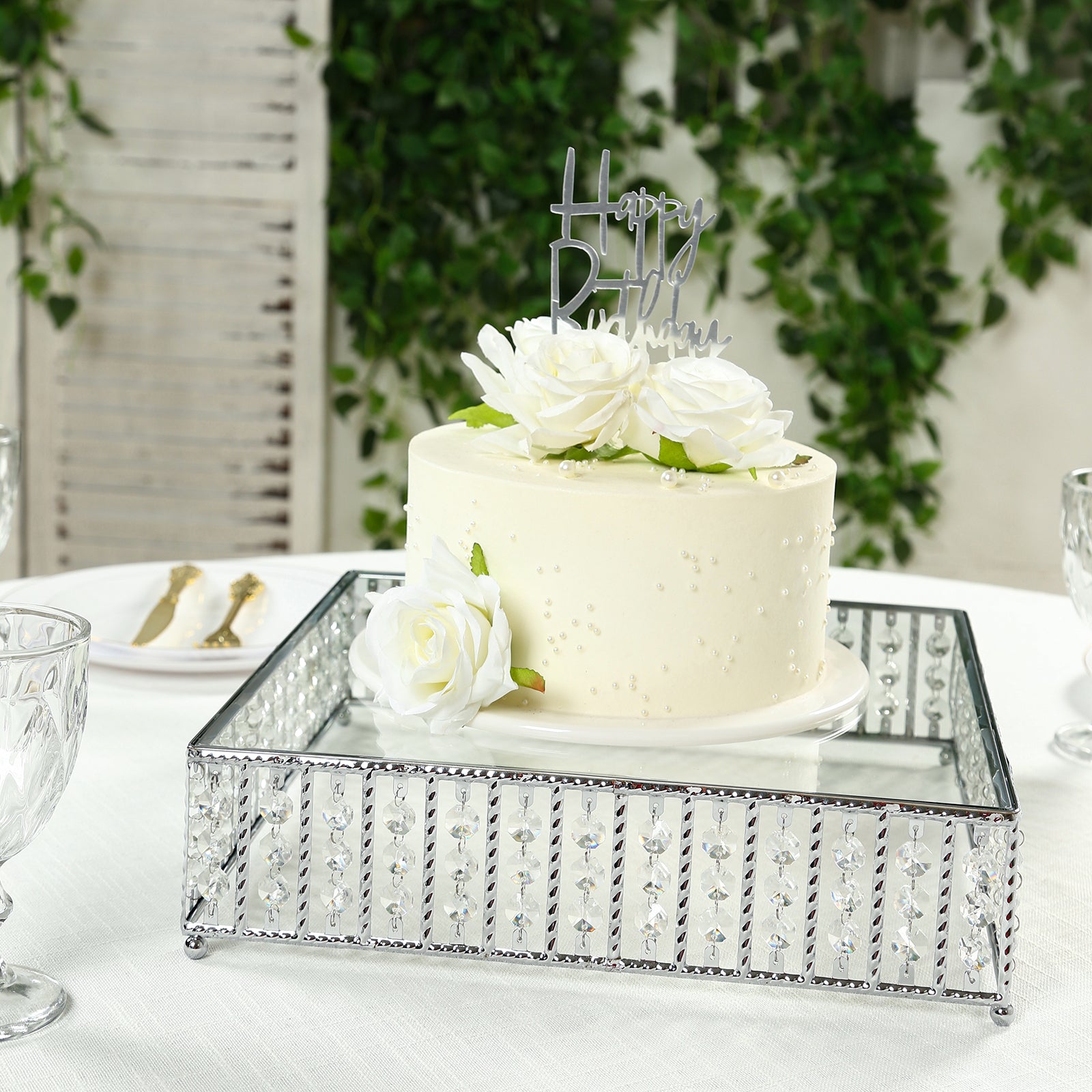 Square silver cake stand with hanging crystal beads and glass top, displaying a cake.