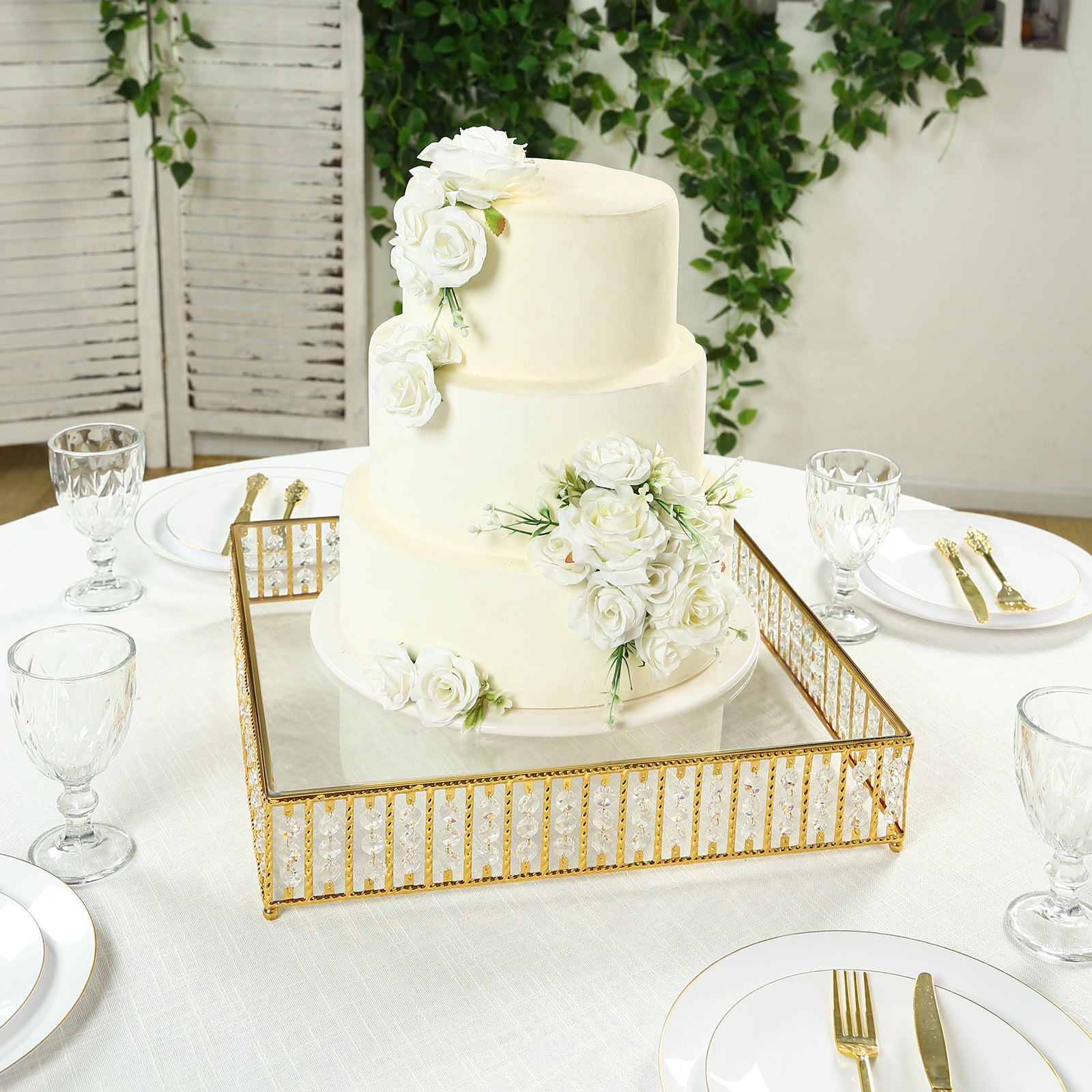 Gold cake stand with hanging crystal beads and glass top, featuring a white floral cake.