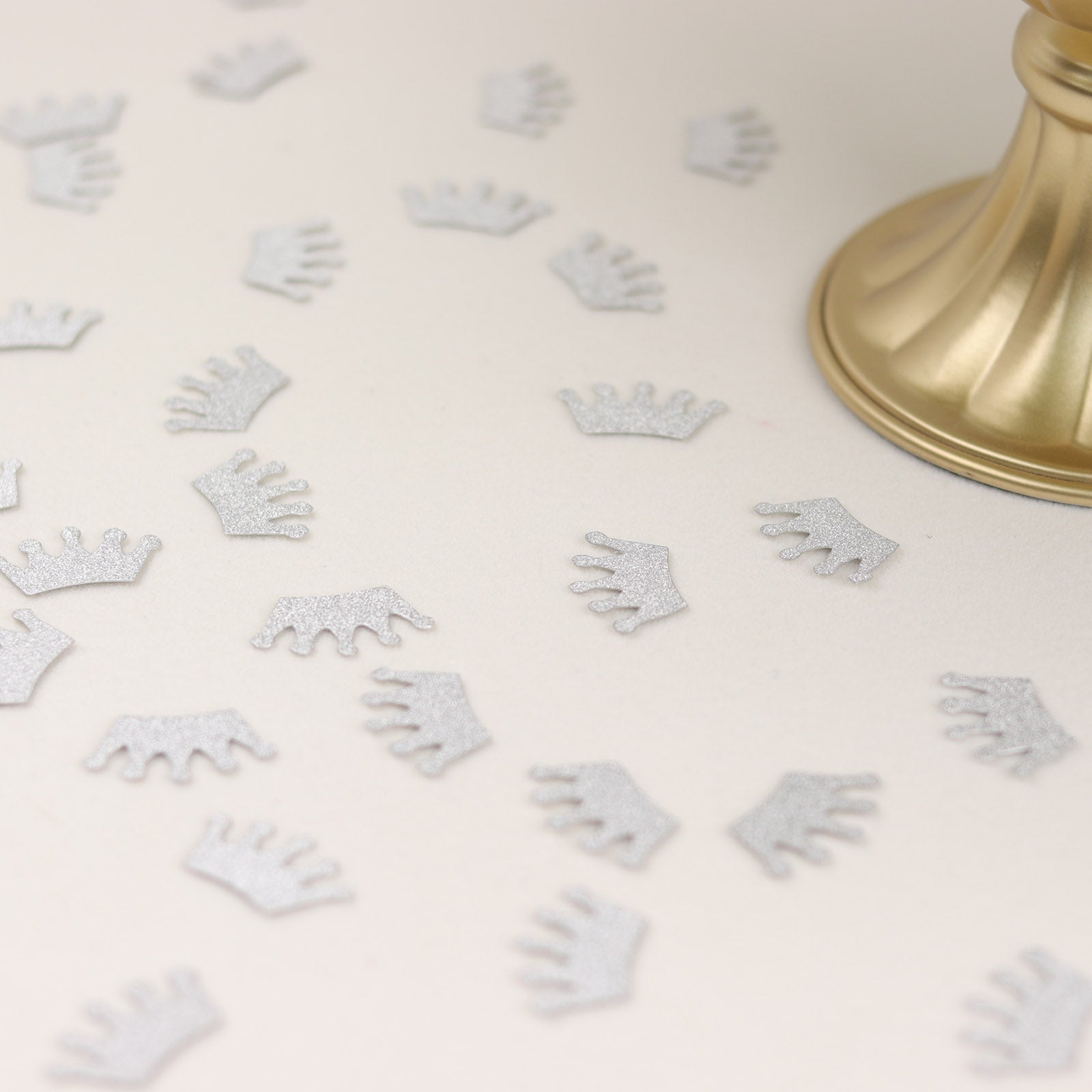 Silver glitter crown confetti scattered on a light background for party decoration.
