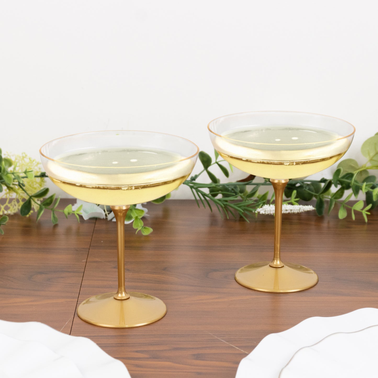 Clear champagne coupes with gold accents on a wooden table with greenery.
