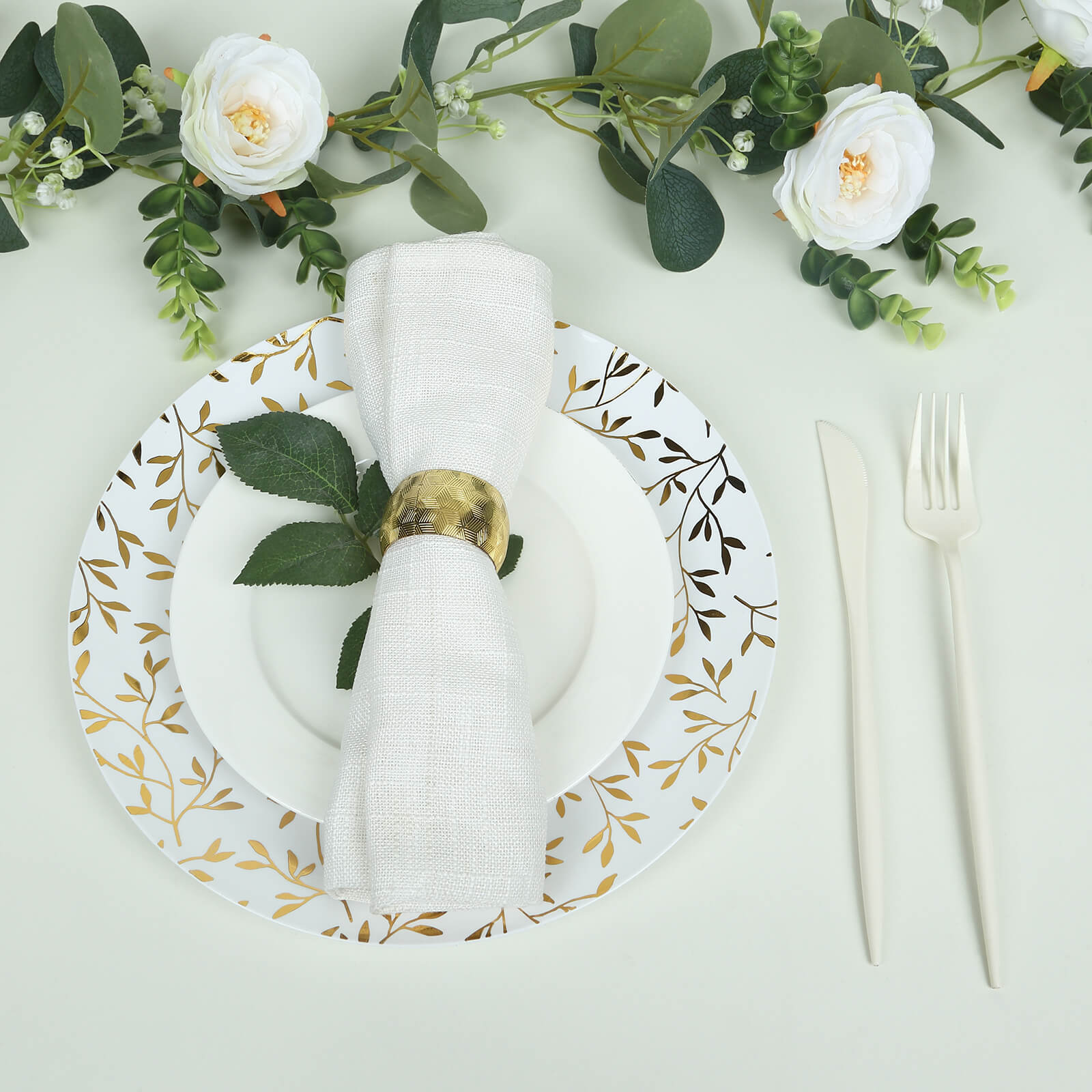 White round plastic party plate with gold leaf design, styled with a napkin and greenery.