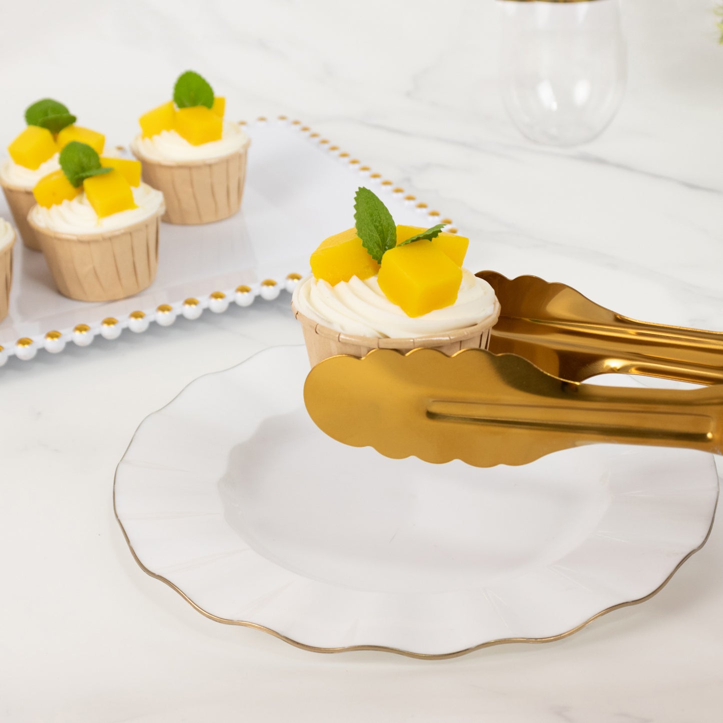 Gold metal kitchen tongs serving cupcakes with mango and mint garnish.