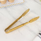 Gold metal kitchen tongs with scalloped edge on a marble surface.