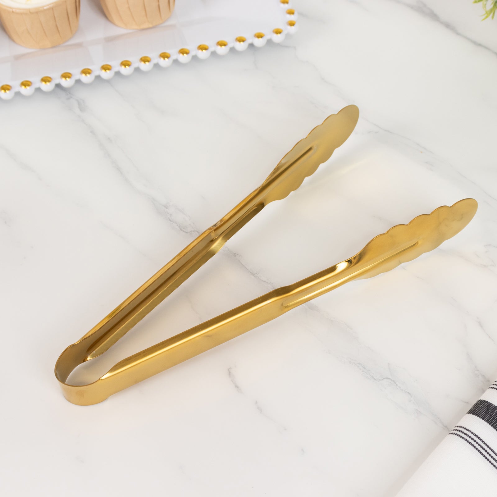 Gold metal kitchen tongs with scalloped edge on a marble surface.
