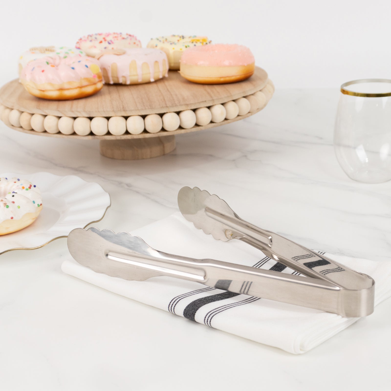 Silver metal kitchen tongs with scalloped edges on a napkin beside a plate of donuts.