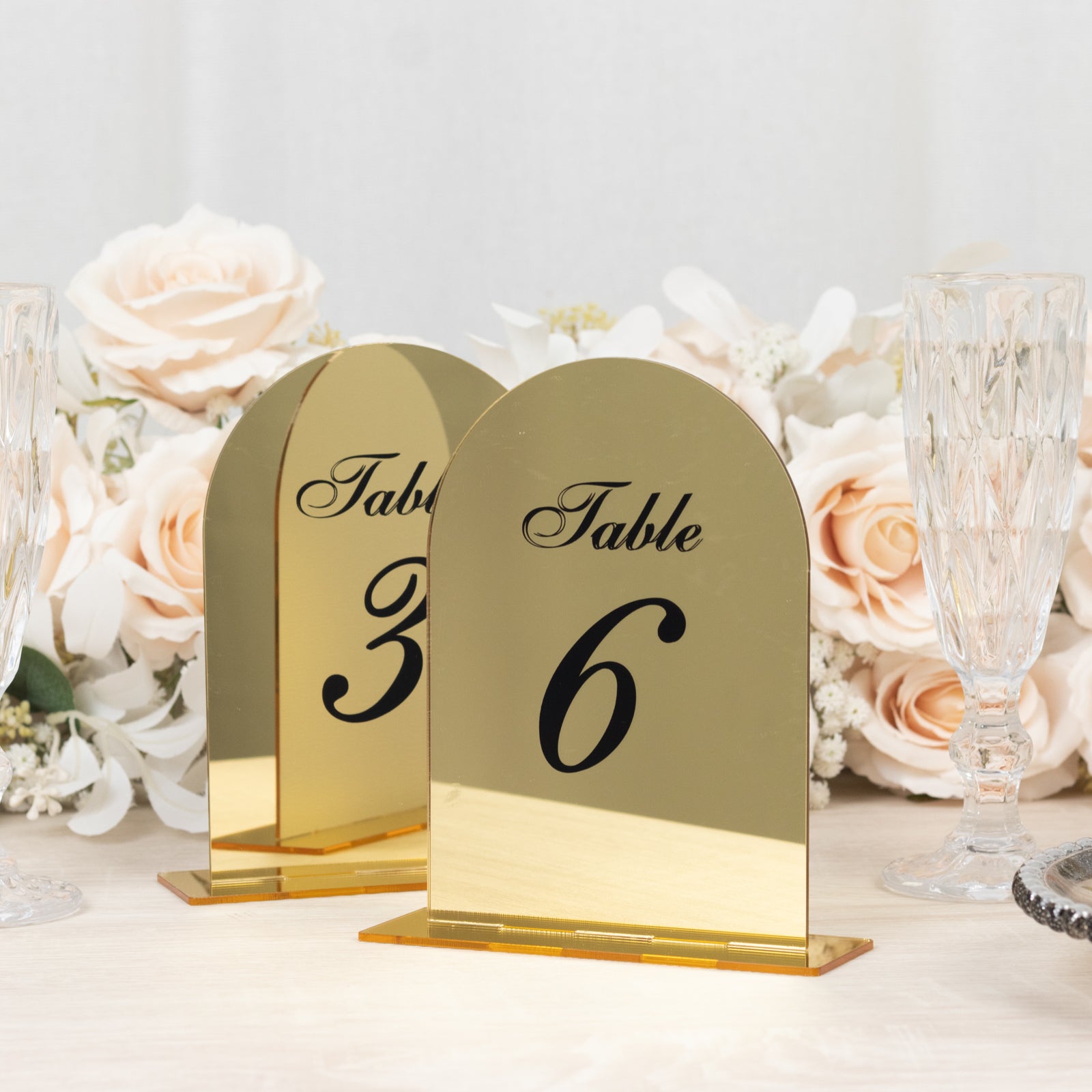 Gold mirror arch table numbers with black print, set against a floral backdrop.
