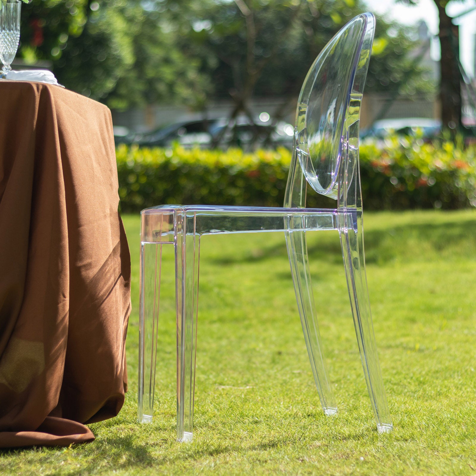 Clear acrylic ghost chair, stackable, with oval back, set outdoors beside a table.