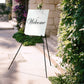 Black metal easel stand holding a welcome sign outdoors.