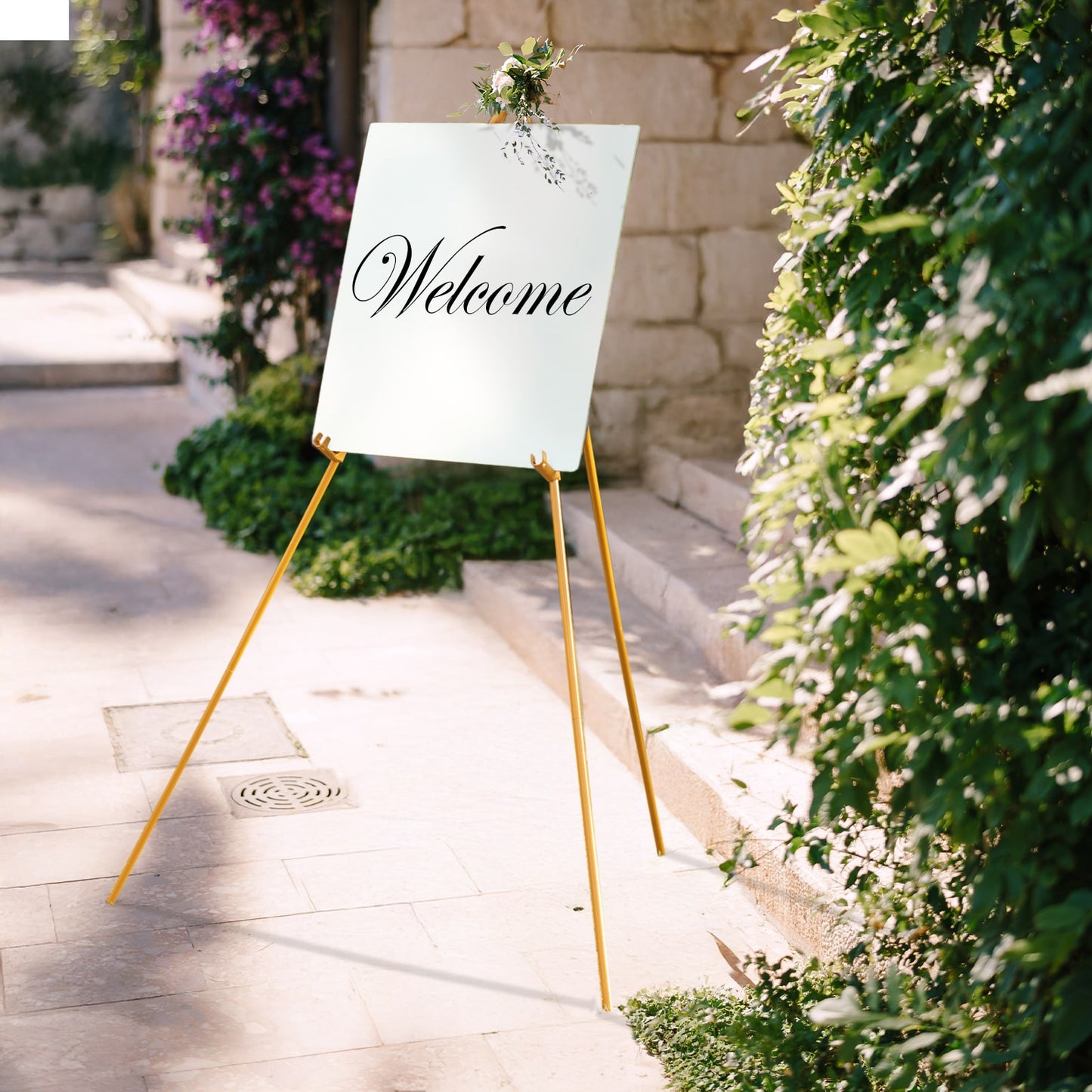 Gold metal easel stand with a welcome sign in a floral outdoor setting.