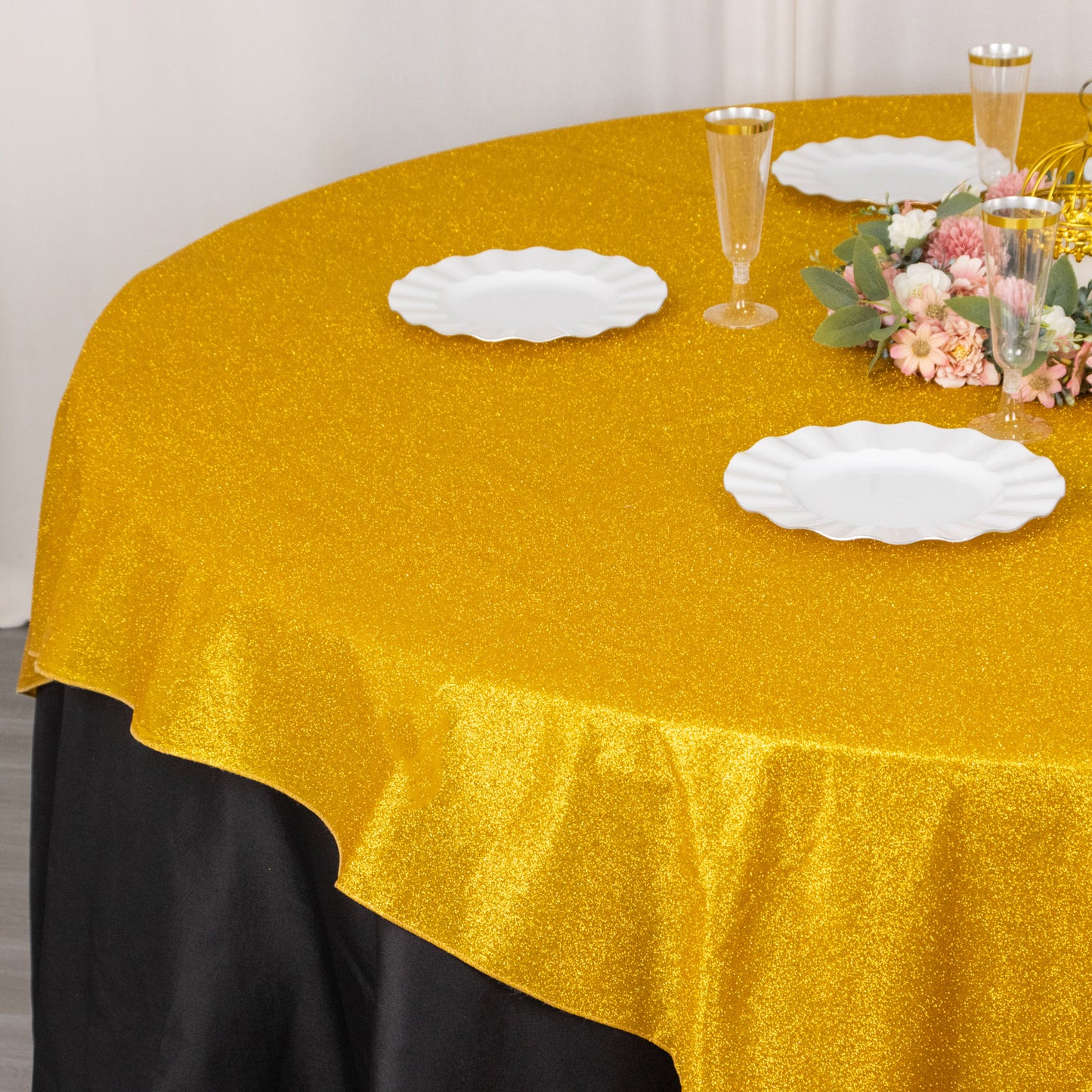 Gold glitter table overlay on a round table with white plates and floral centerpiece.