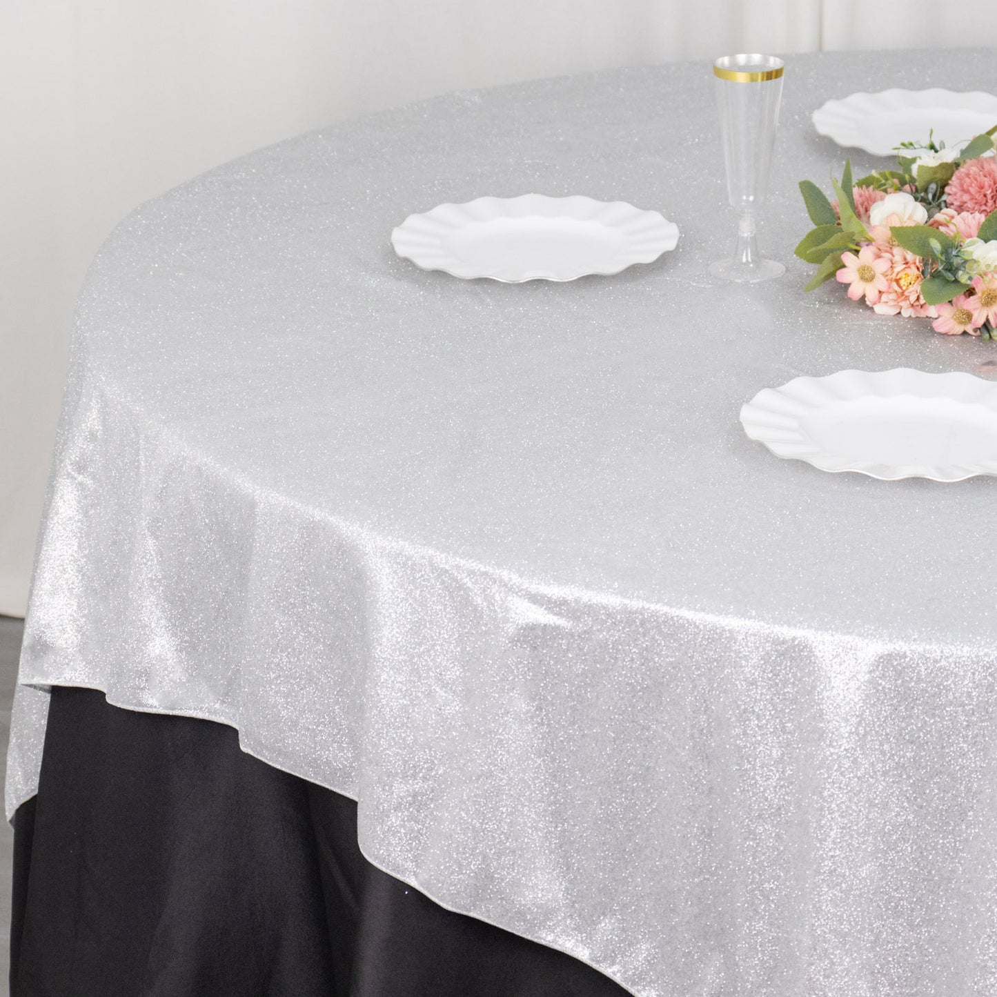 Silver glitter table overlay on a round table with white plates and floral arrangement.