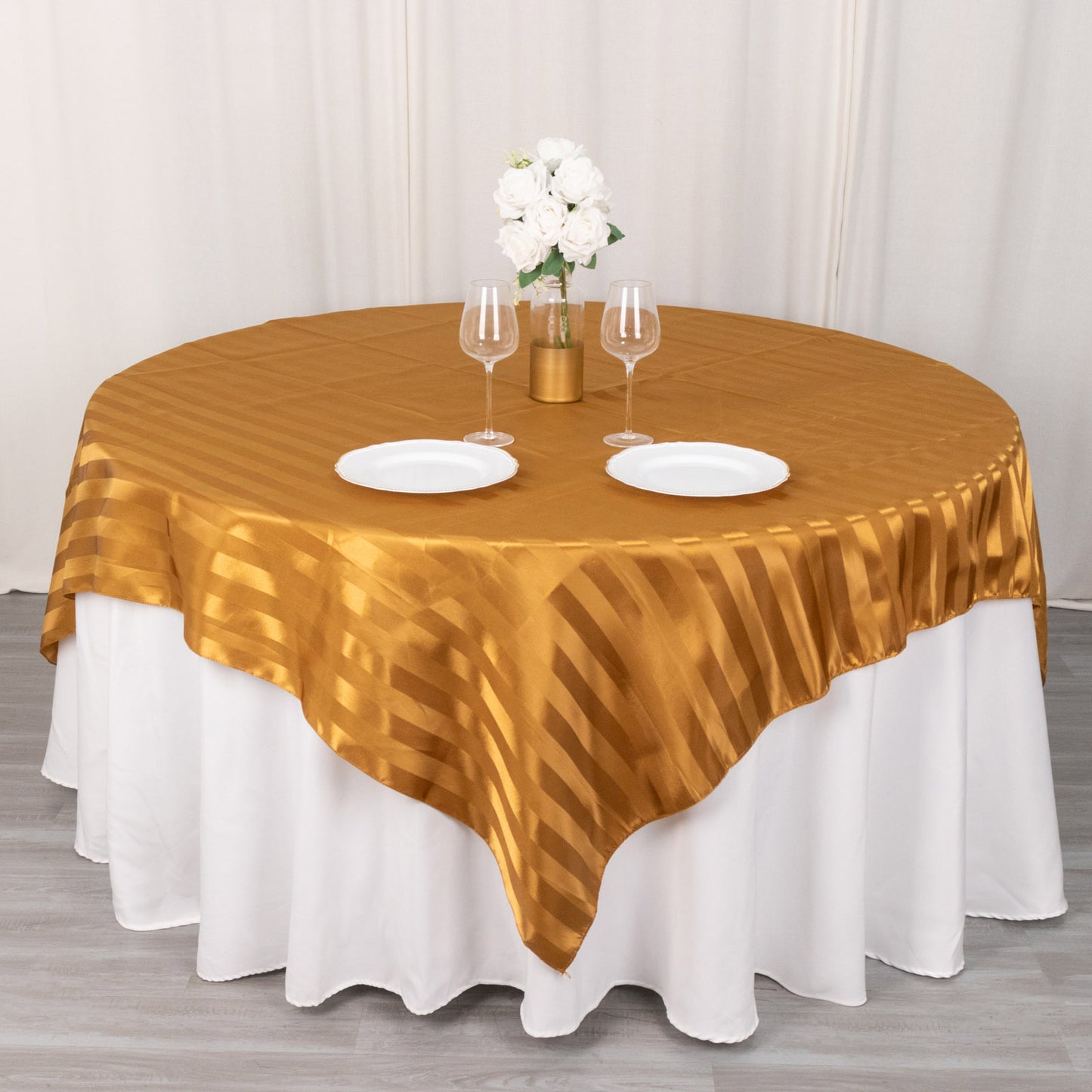 Gold satin stripe table overlay on a round table with white plates and flowers.