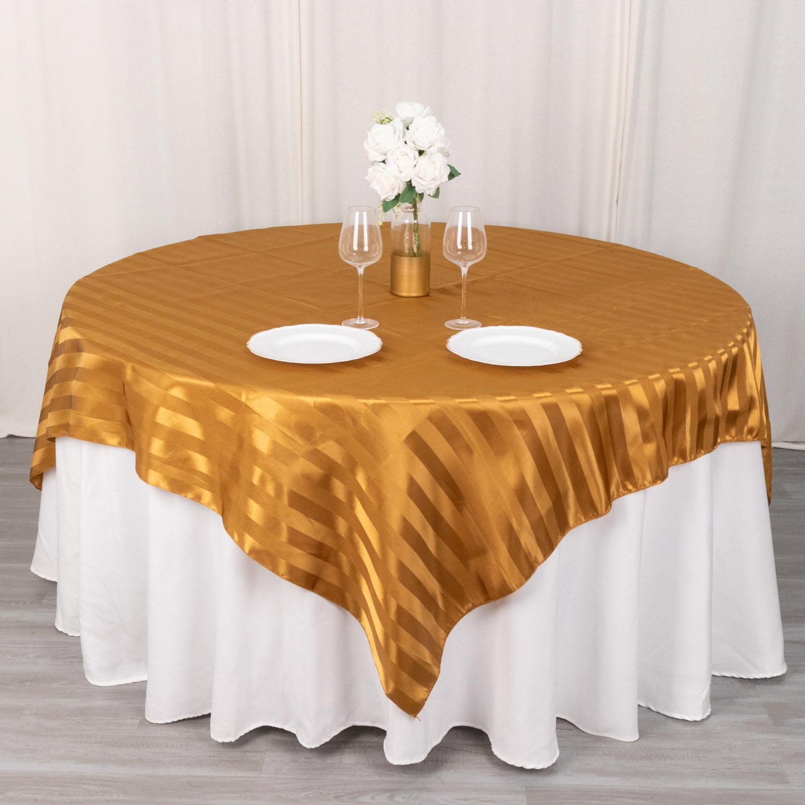 Gold satin stripe table overlay on a round table with white plates and flowers.