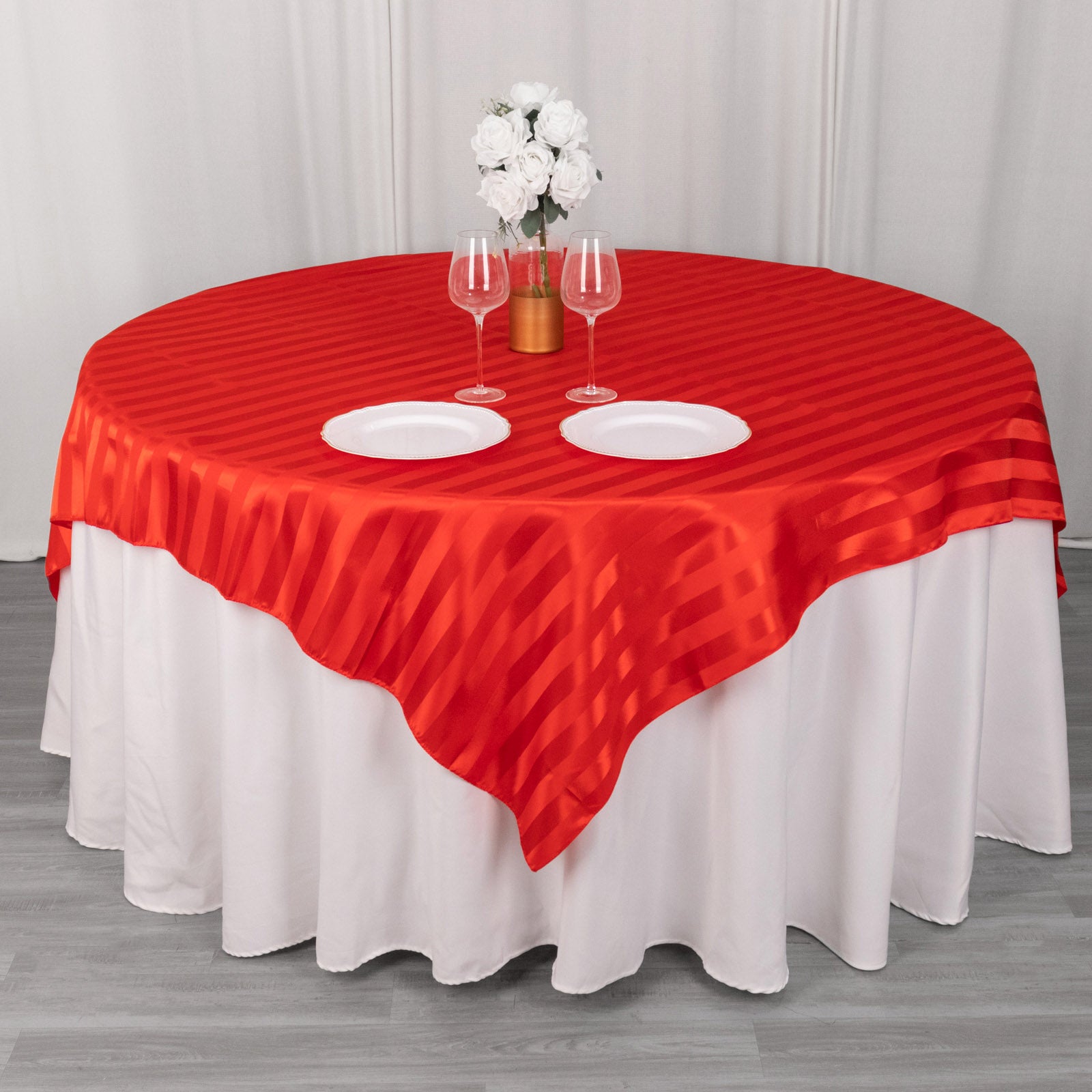 Red satin stripe table overlay on a round table with white plates and flowers.