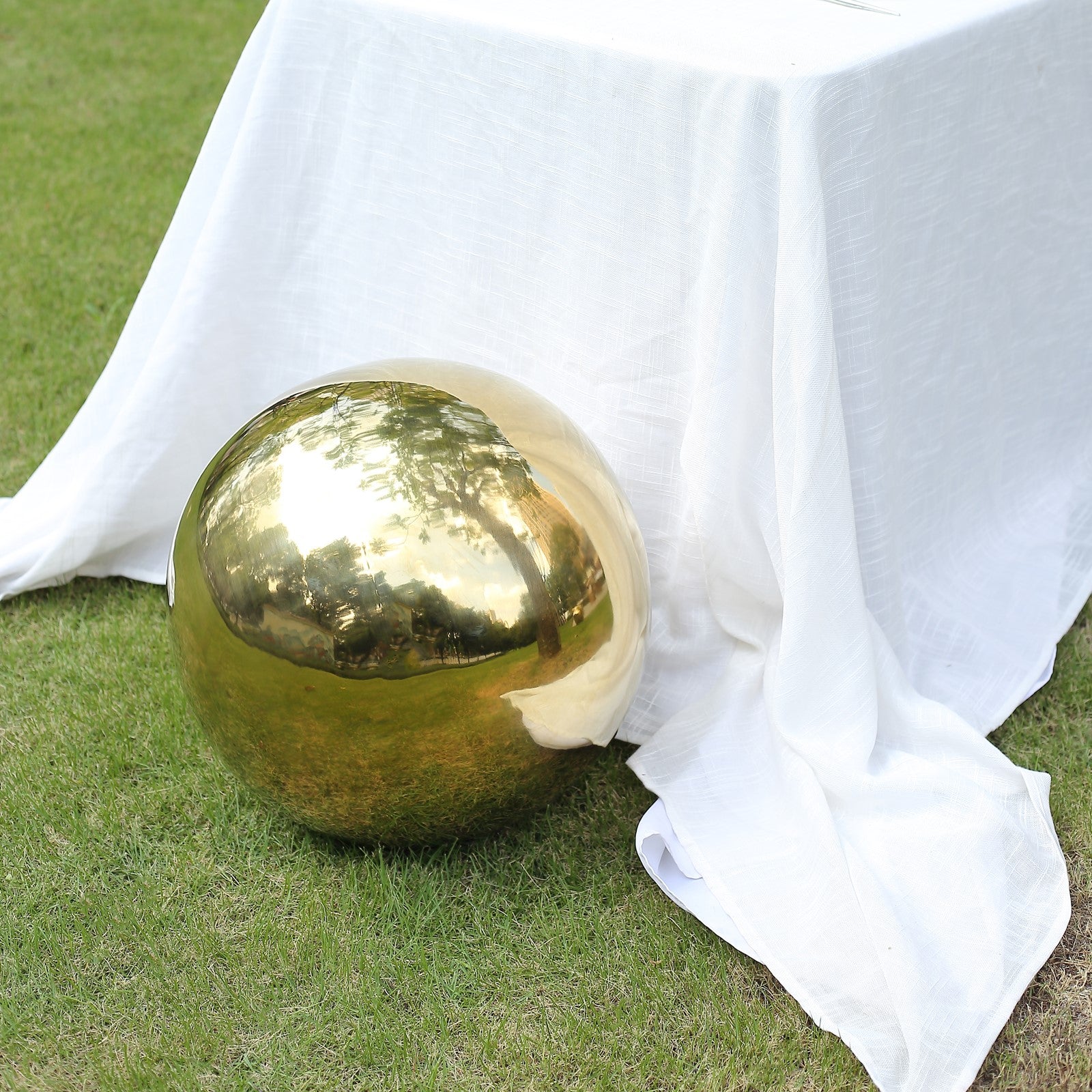 Gold shiny mirror gazing ball resting on grass beside a white tablecloth.