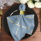 Modern dusty blue and geometric gold dinner napkin on a black plate.