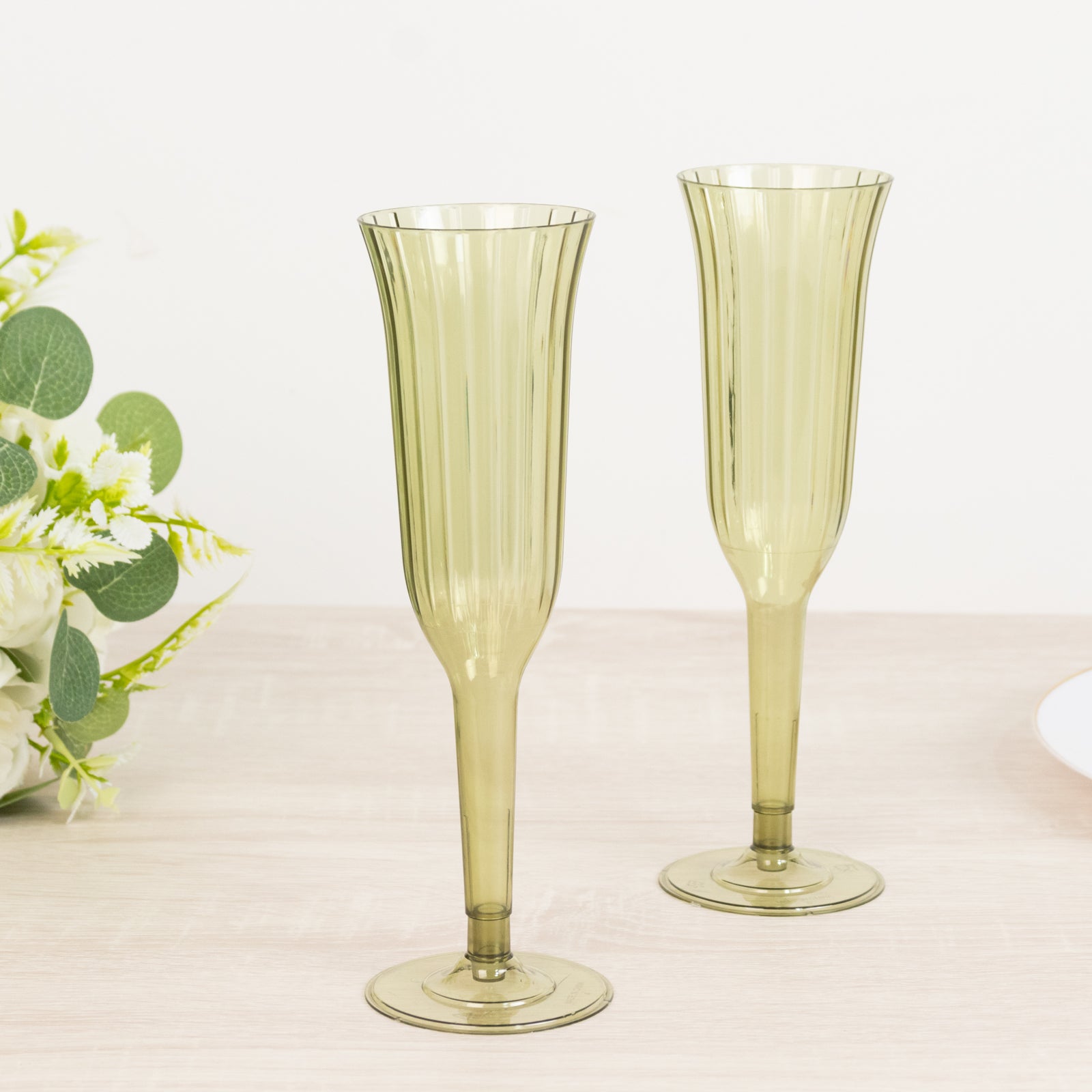 Dusty sage green plastic champagne flutes on a table with flowers.