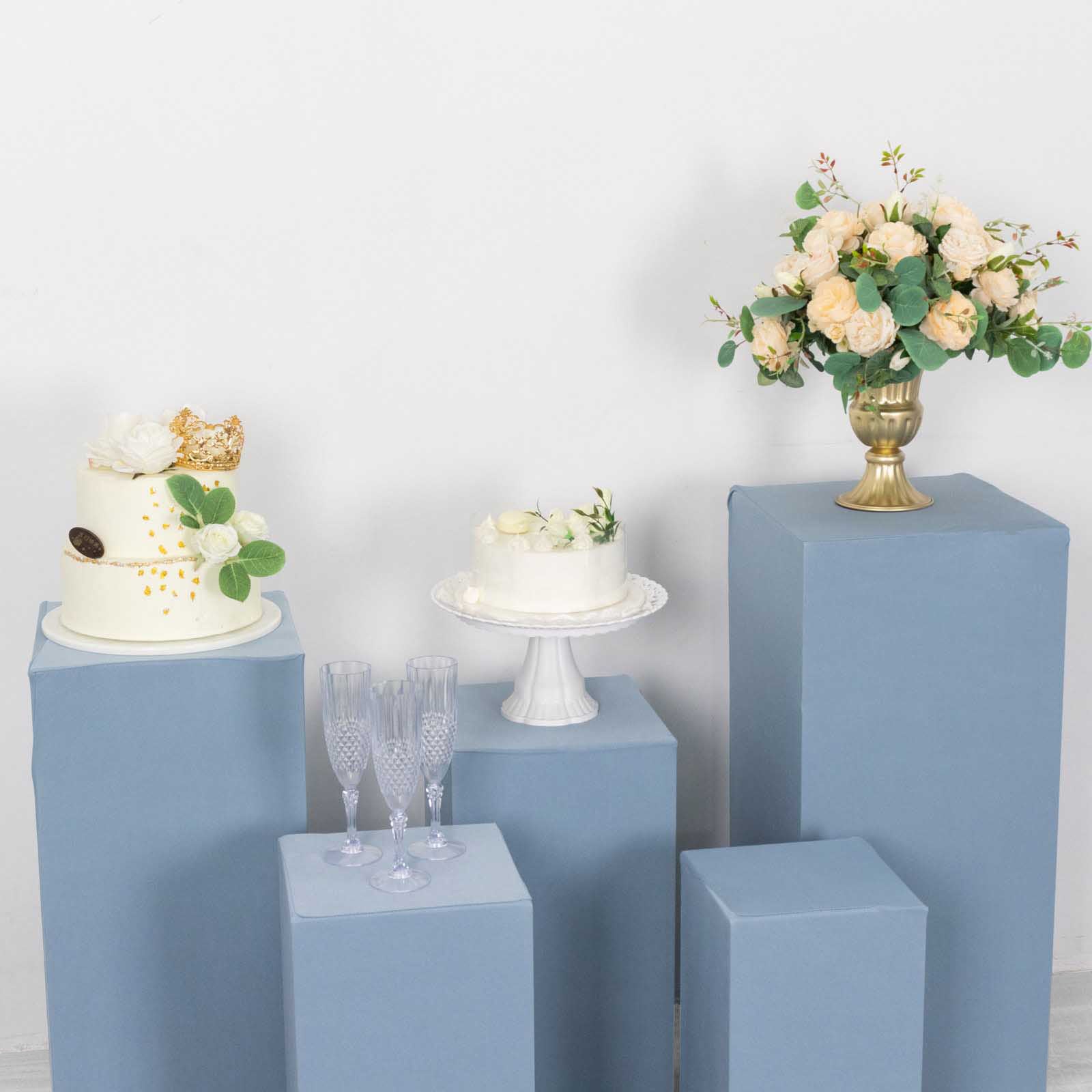 Dusty blue spandex pedestal stand covers with cakes and flowers displayed.