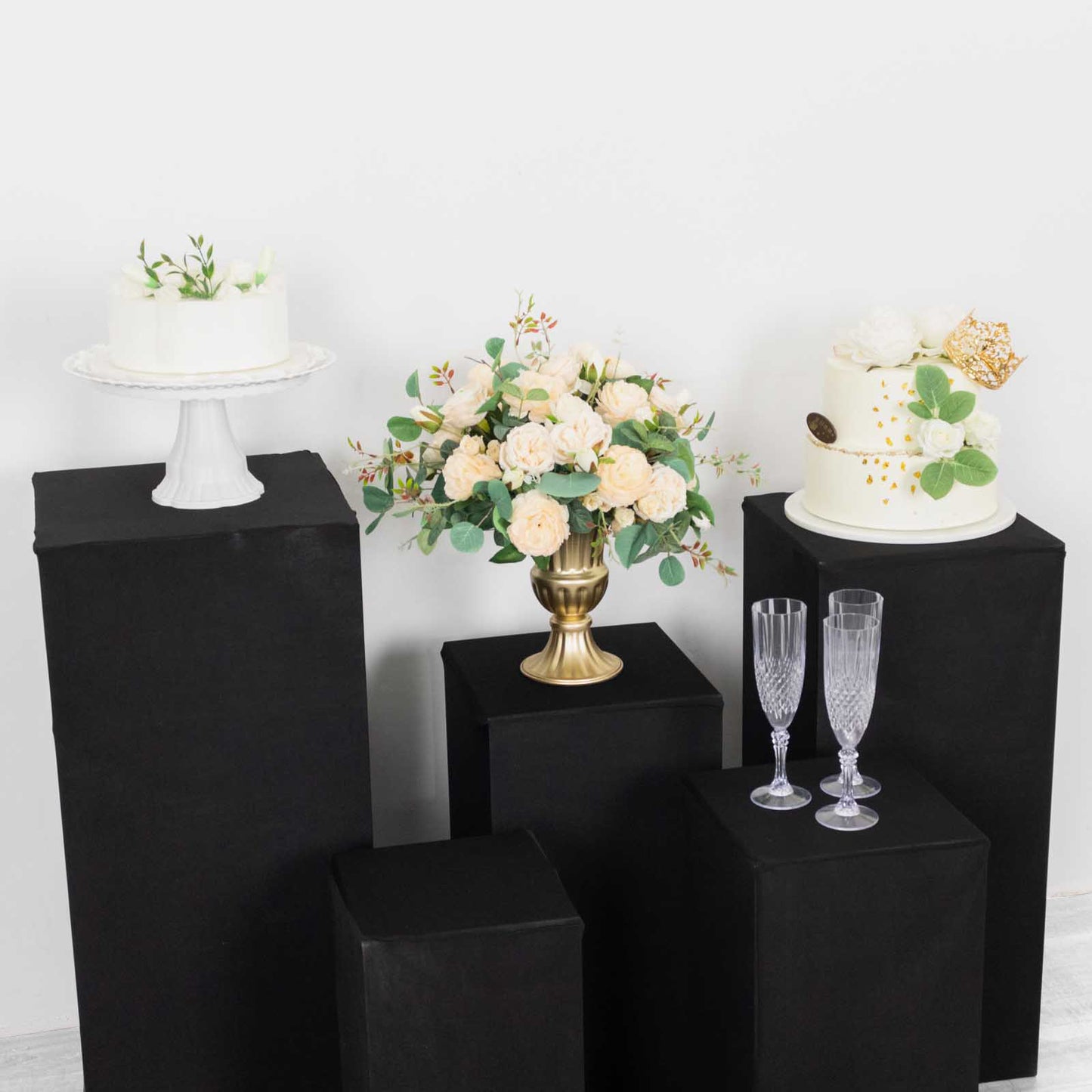 Black spandex pedestal stand covers on display with floral arrangements and cakes.