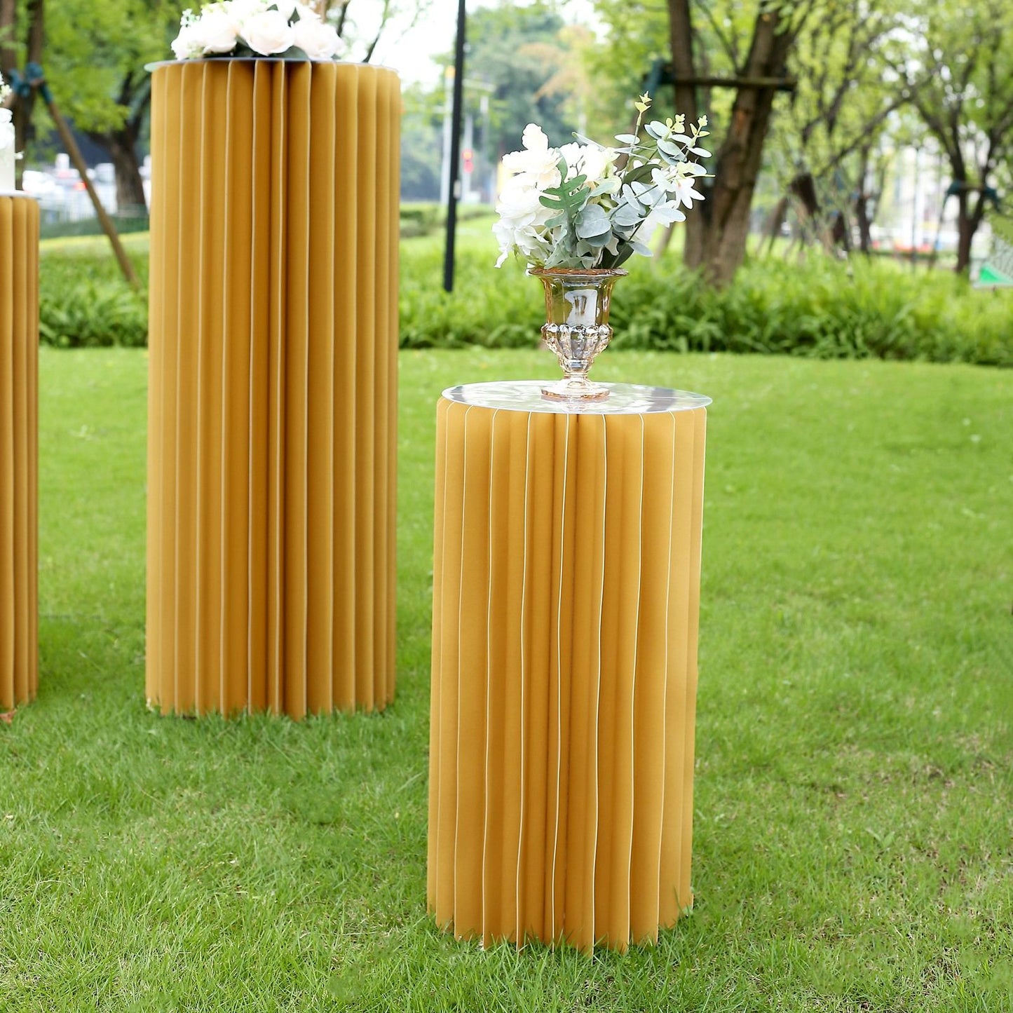 Gold cylinder display column stand with top plate in a garden setting.