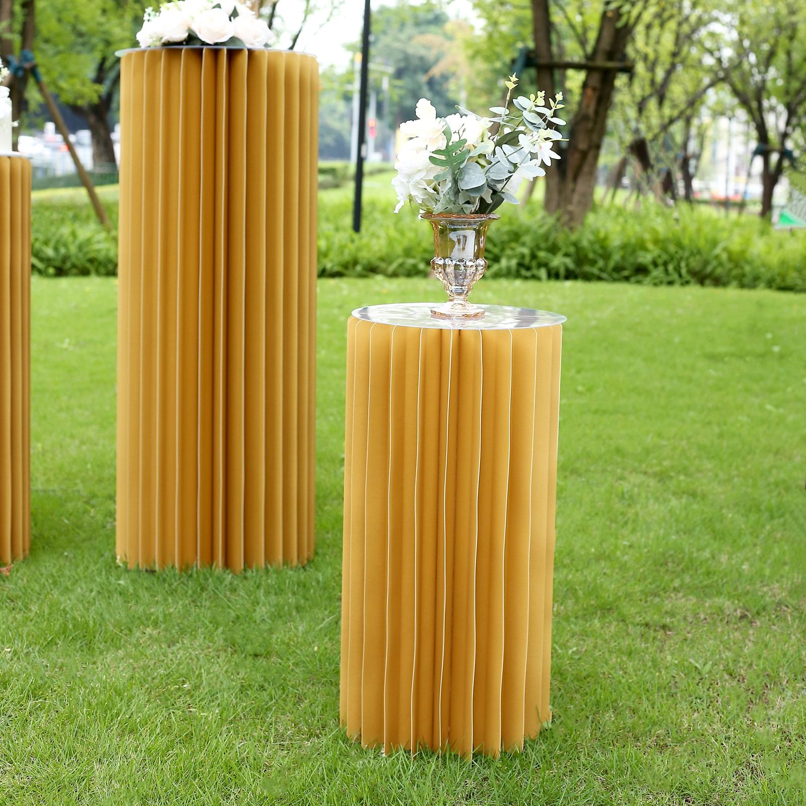 Gold cylinder display column stand with top plate in a garden setting.