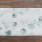 White table runner with green eucalyptus leaves print on a wooden surface.