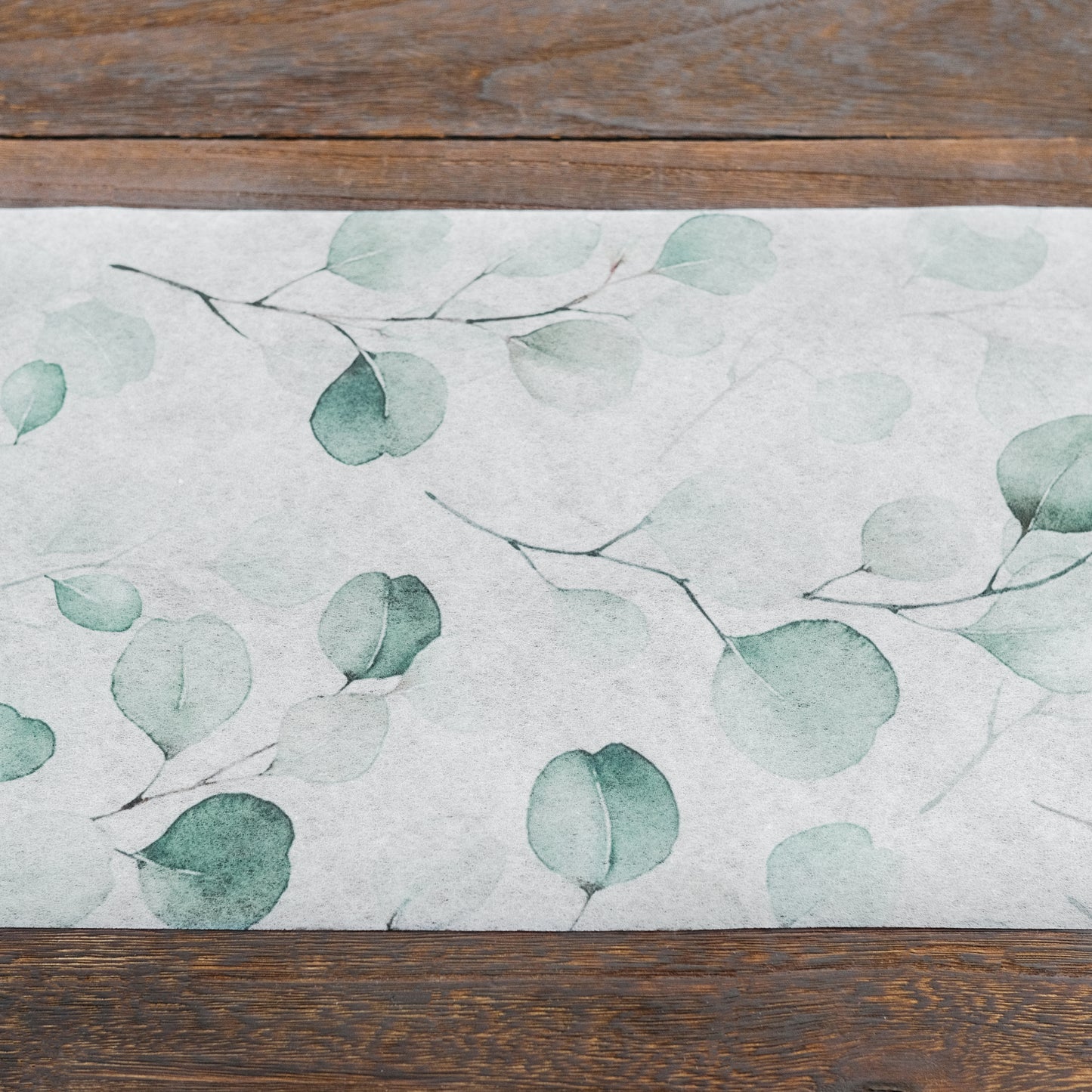 White table runner with green eucalyptus leaves print on a wooden surface.
