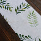 White table runner with green olive leaves print on a wooden surface.